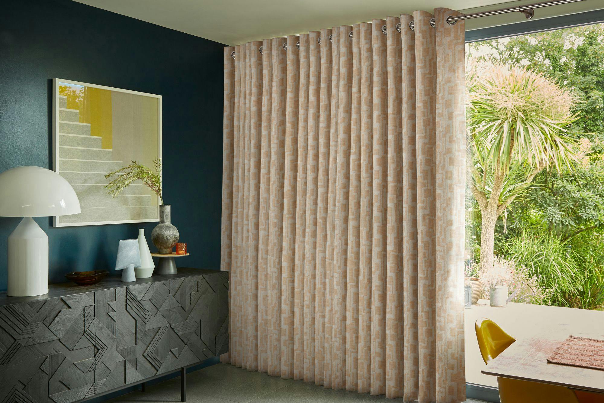 Patterned curtains hang beside a large window overlooking a garden. A dark, geometric-textured cabinet holds decorative vases, a lamp, and a framed artwork under a teal wall.