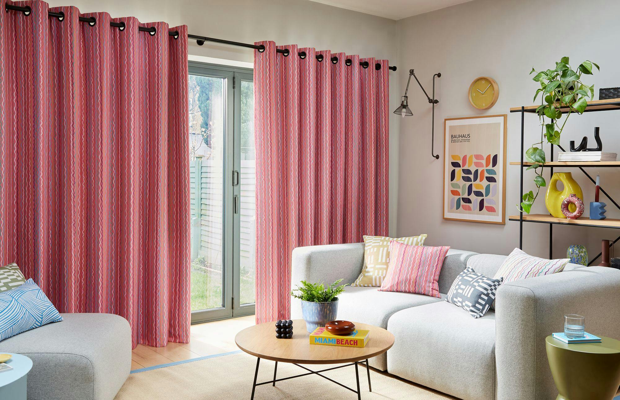 Curtains hang in a living room, partially covering sliding glass doors. The room has a light gray sofa, colorful pillows, a wooden table with books "MIAMI BEACH," and plants on shelves. A framed poster reads "BAUHAUS."