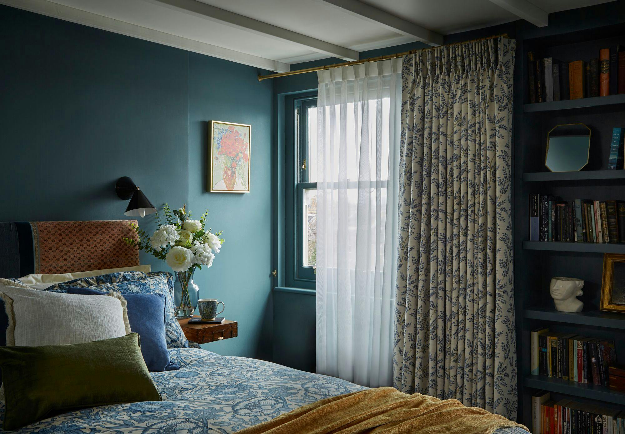 A bedroom with a patterned bed, floral curtains, and a vase of flowers on a side table. Books and a decorative sculpture fill a nearby shelf, enhancing the cozy atmosphere.