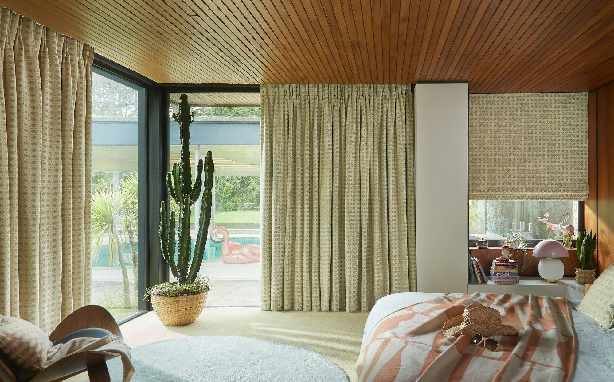 Bed with patterned blanket and a hat lies in a room with wood-paneled ceiling, large cactus by window, and view of garden with inflatable flamingo by pool. Books and lamp on shelf.