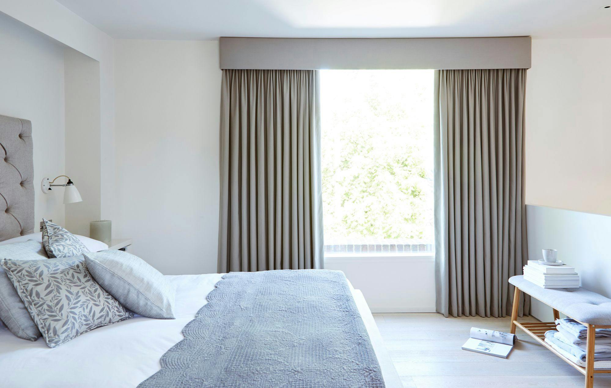 A neatly made bed with patterned pillows and a gray throw blanket is alongside a window with closed beige curtains. A bench holds stacked books, a cup, and magazines nearby.