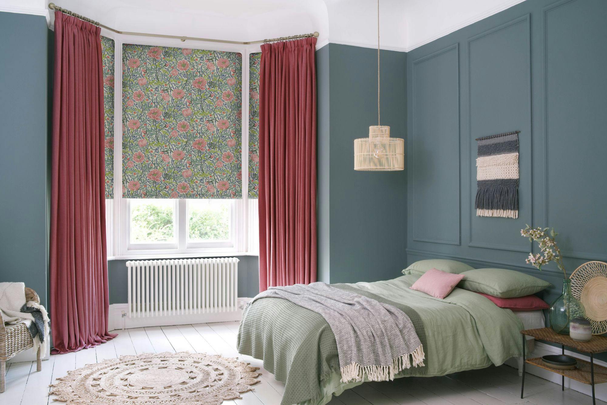 A bedroom features a bed with green linens and a gray throw. Pink curtains frame a window with floral blinds. Decorative elements include a woven rug, pendant light, and wall hanging.