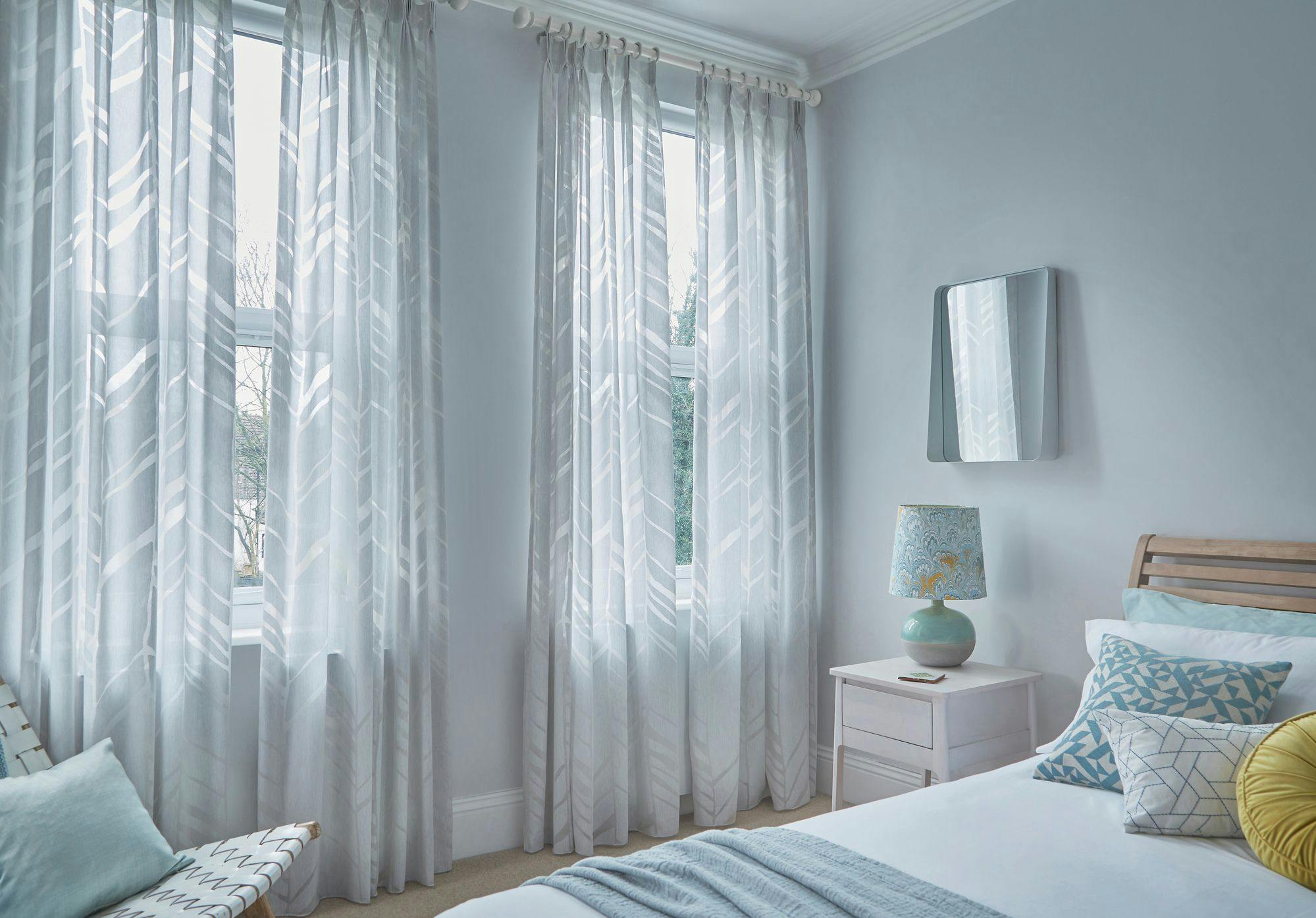 Curtains hang from two large windows, filtering sunlight into a softly lit bedroom featuring a neatly made bed with patterned cushions, a bedside table with a decorative lamp, and pale blue walls.