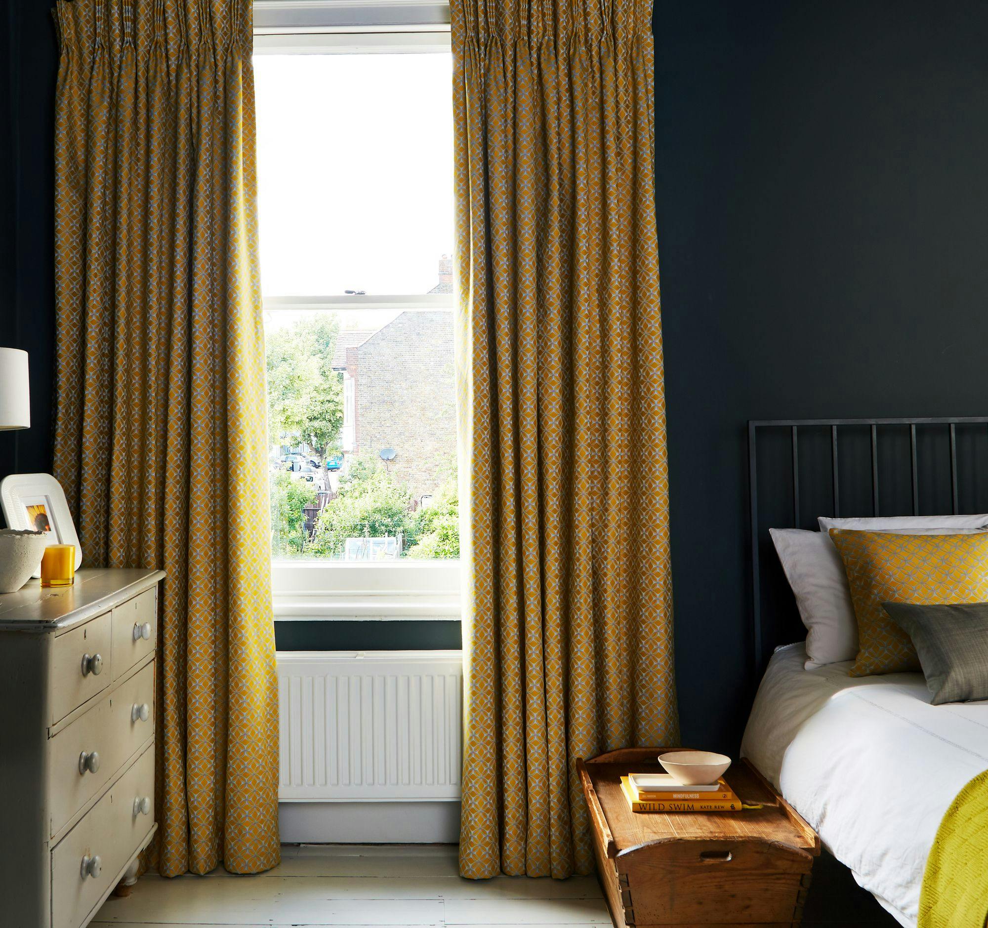 Yellow patterned curtains hang beside a window, framing views of a garden and buildings outside. In the room, a bed with pillows and a dresser with a lamp are visible.
