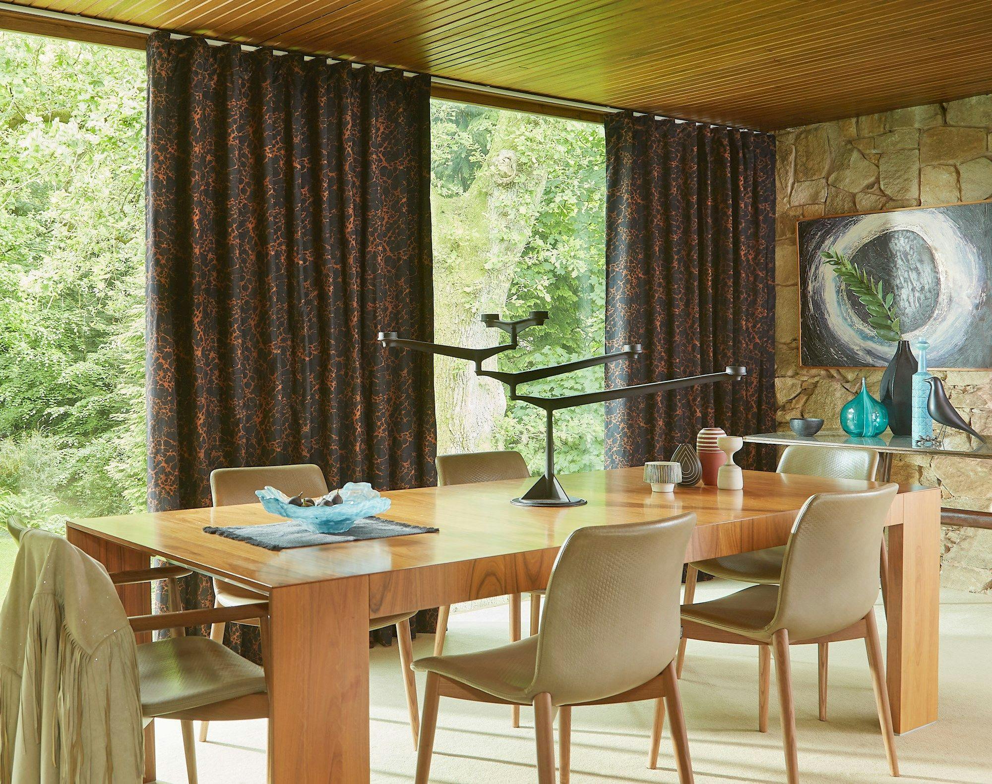 A wooden dining table with a modern lamp and decorative items sits in a room with stone walls, surrounded by chairs and large windows with dark curtains overlooking greenery.