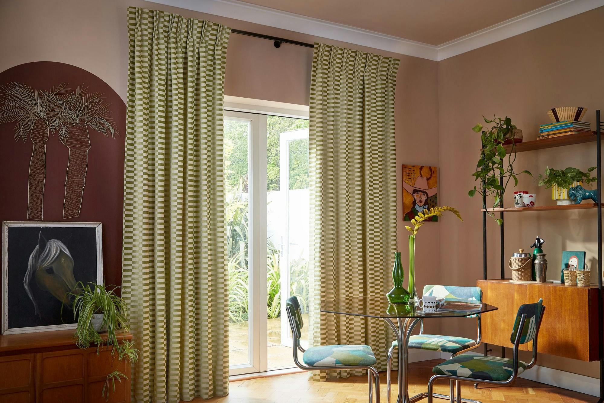 Curtains hang beside a glass door, partially open, revealing a garden. A round glass table with chairs and a vase sits nearby, surrounded by plants, shelves, and artwork in a cozy room.