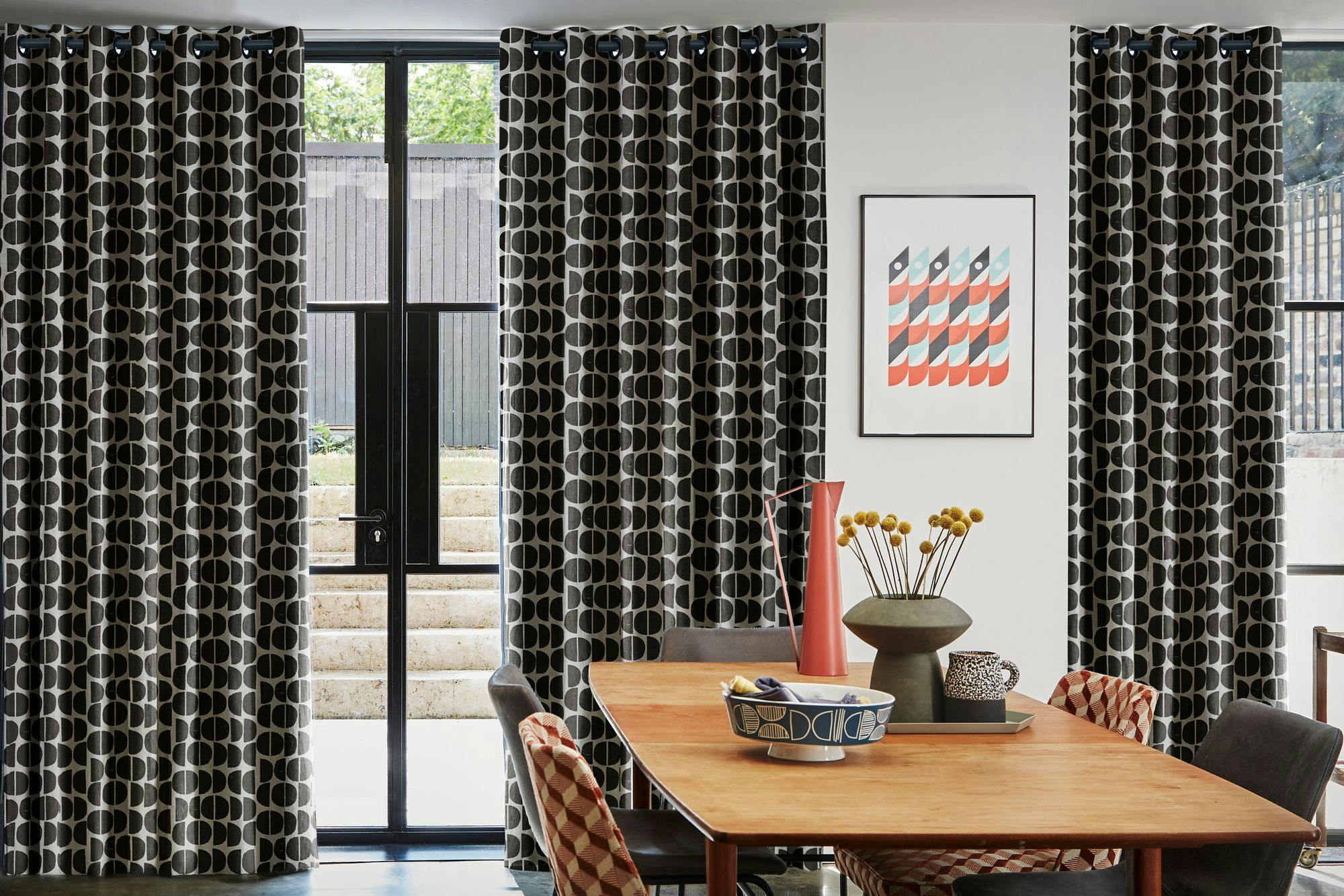 Curtains with a black and white geometric pattern hang around a glass door. A wooden table holds ceramic vases and a decorative bowl, set in a modern room with abstract wall art.
