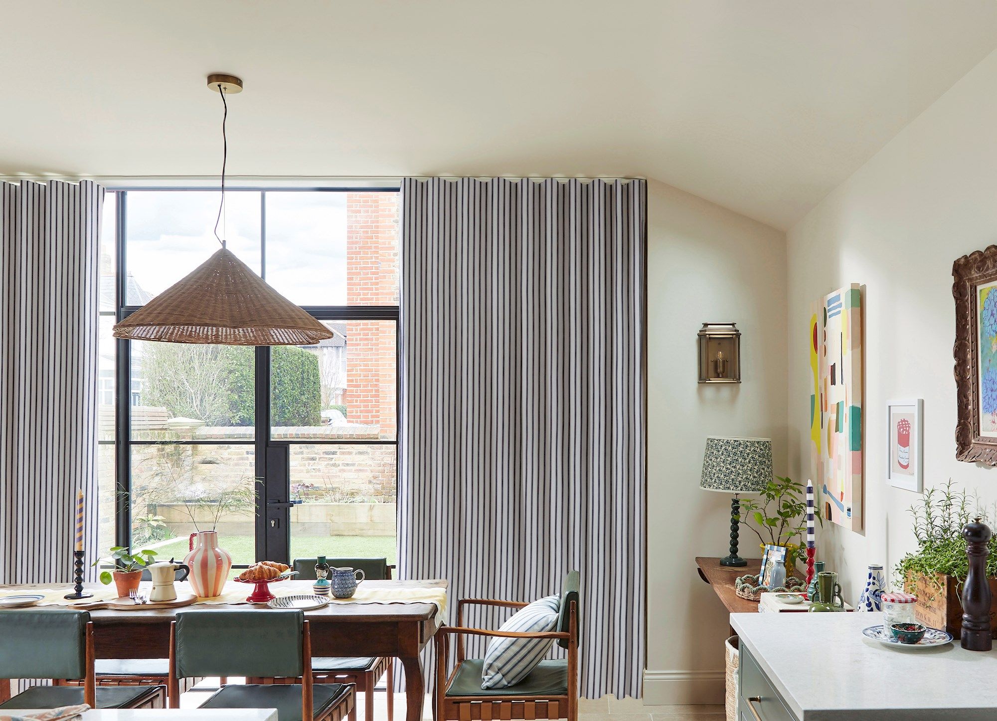 Dining table set with ceramics and plants, beneath a wicker pendant light. Large striped curtains cover a windowed wall, revealing a brick exterior beyond. Art decorates the adjacent wall.