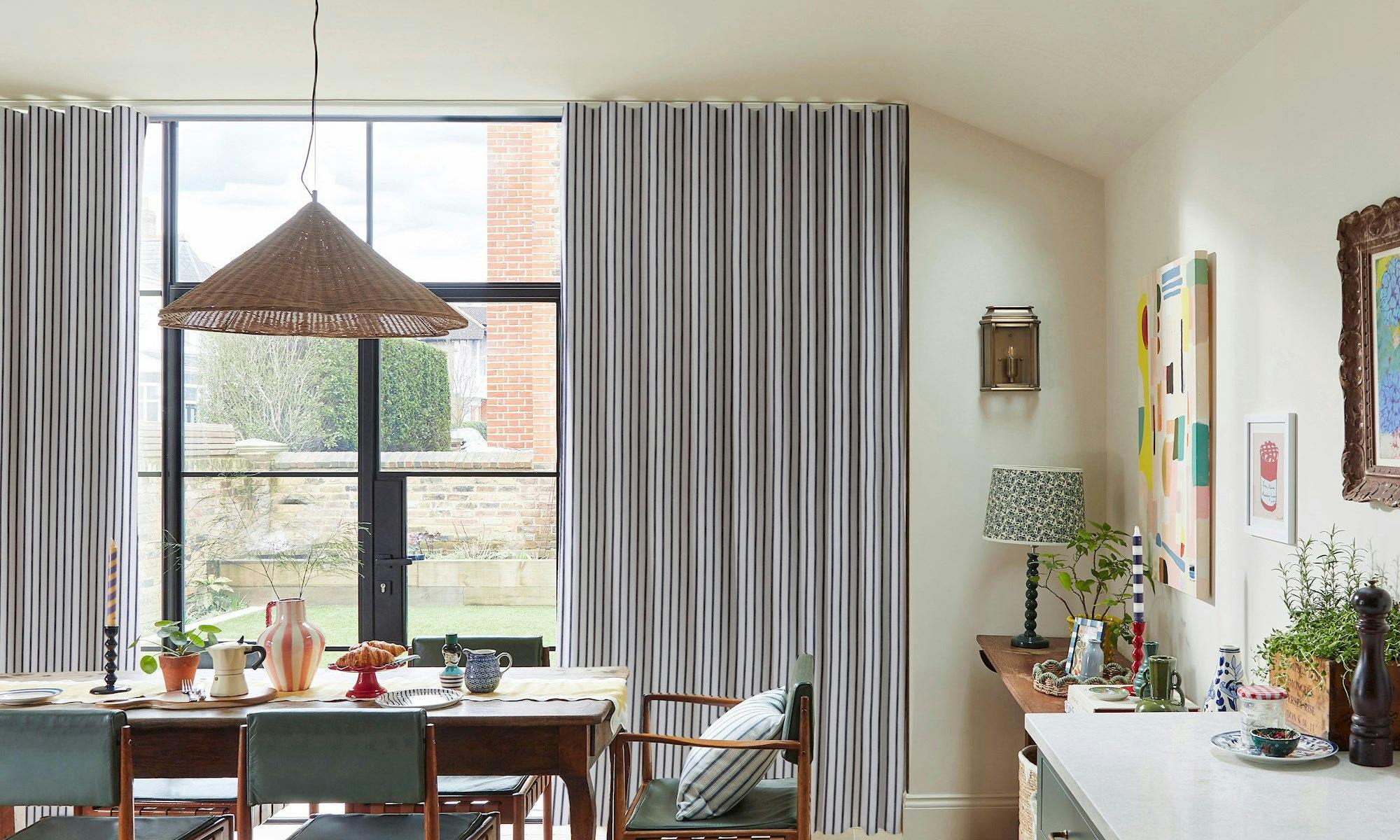 Dining table set with ceramics and plants, beneath a wicker pendant light. Large striped curtains cover a windowed wall, revealing a brick exterior beyond. Art decorates the adjacent wall.