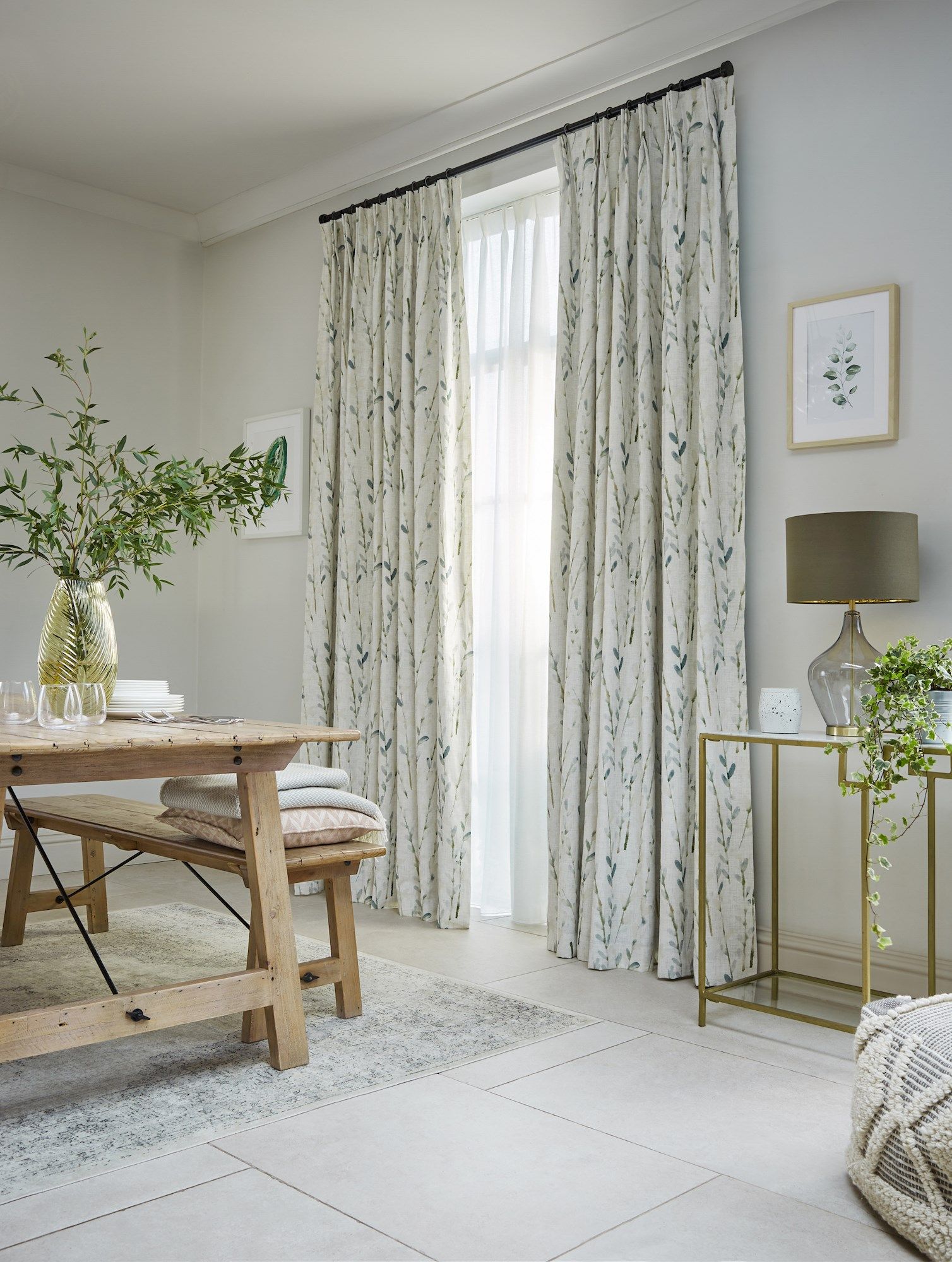 Curtains cover a large window, adorned with a delicate leaf pattern. A wooden dining table with plates and glasses sits nearby, surrounded by a light, airy living room setting.