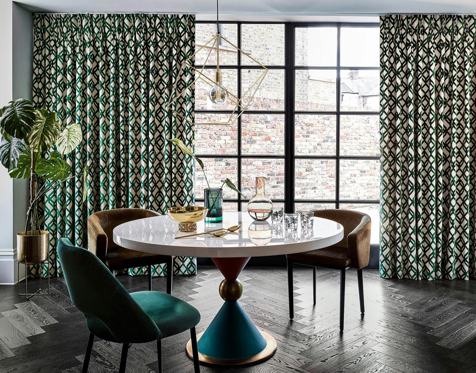 A round table with decorative items sits in front of patterned curtains and a large window in a modern, stylish room with dark wood flooring and green upholstery.