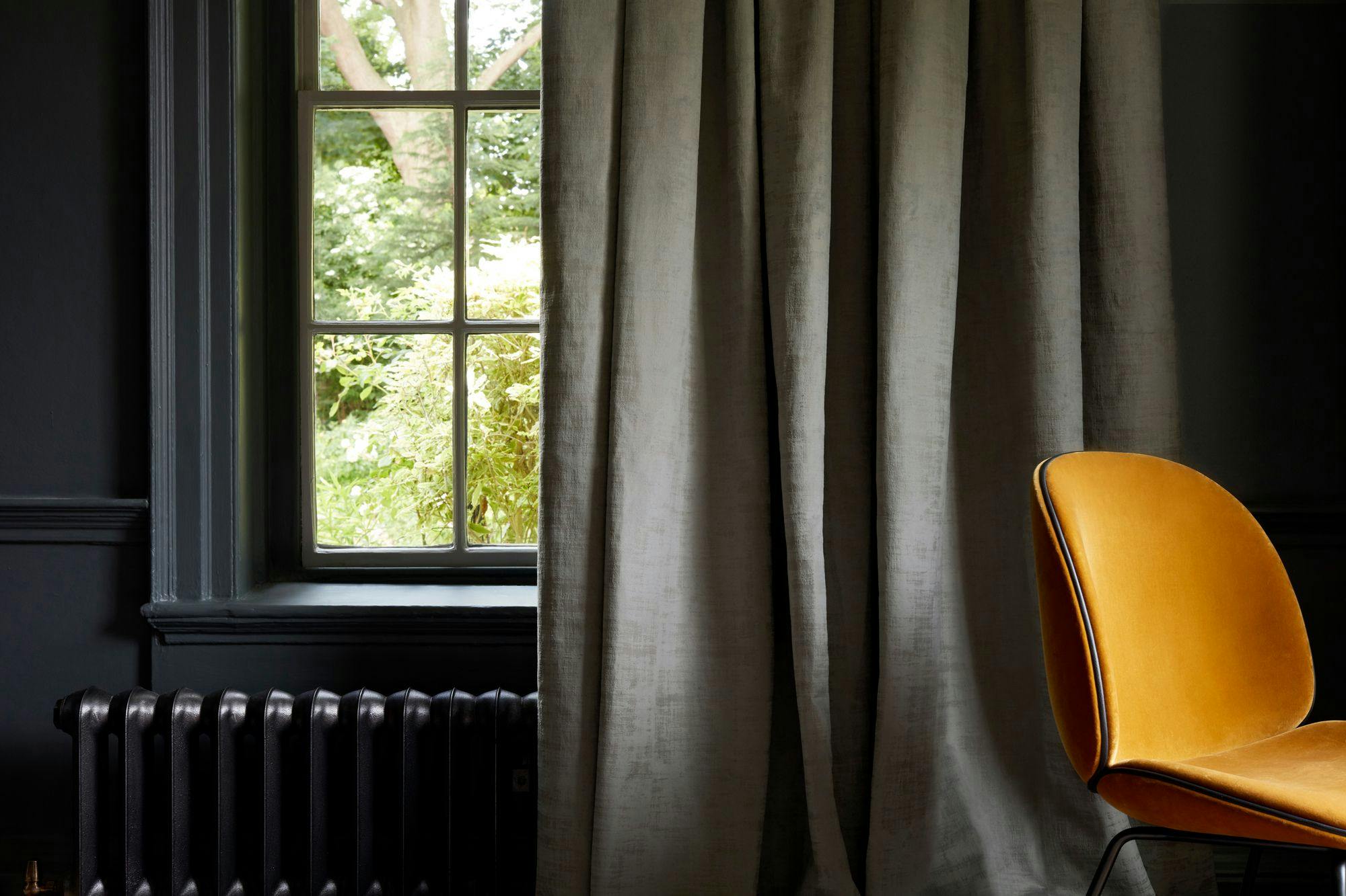 A mustard-yellow chair sits in front of heavy gray curtains, beside a window showcasing lush greenery outside. A dark radiator is positioned below the window, highlighting a cozy interior setting.