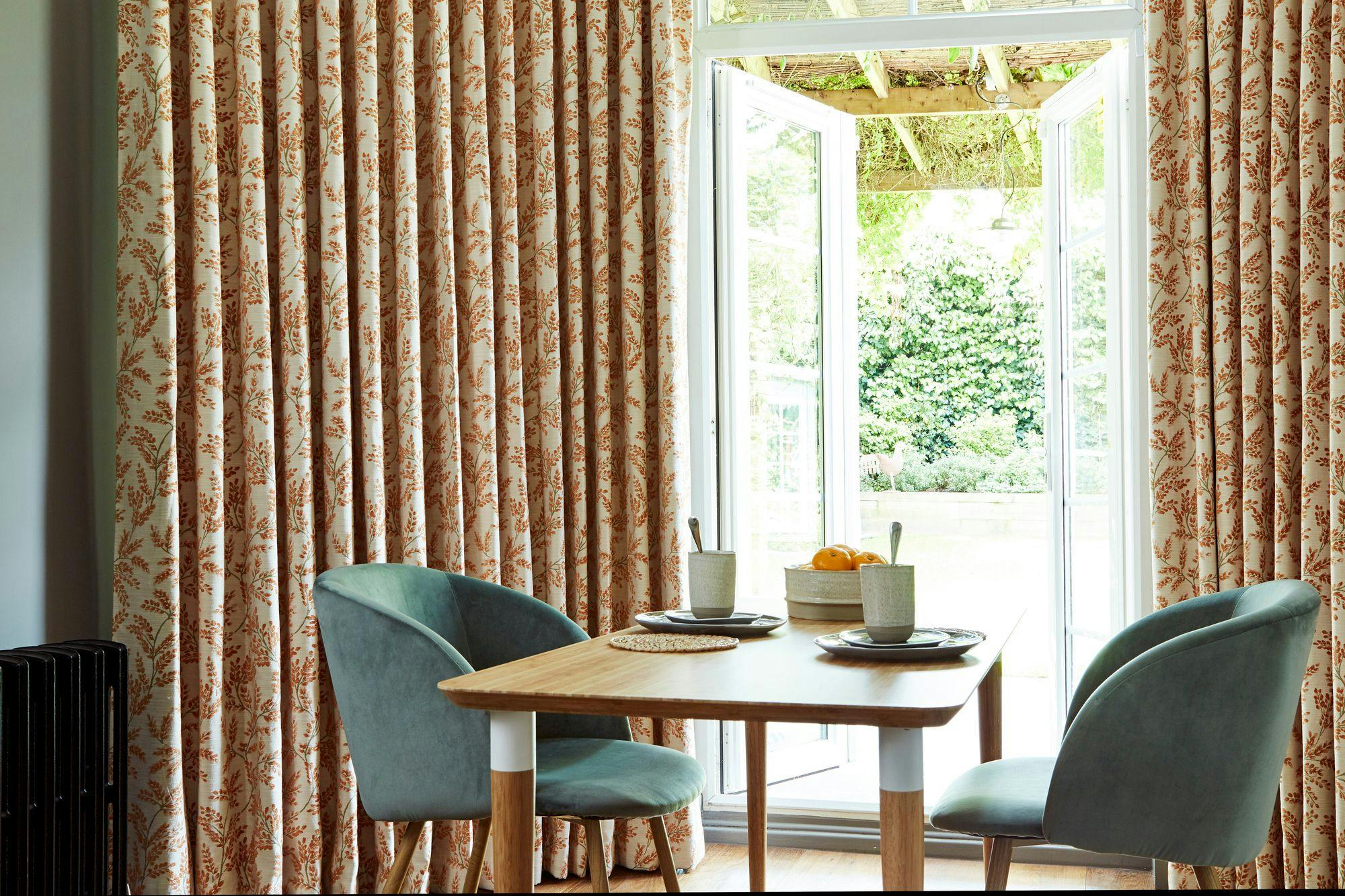 A wooden table with breakfast items sits between two green chairs beside floral patterned curtains. Open glass doors reveal a sunny, lush garden outside.
