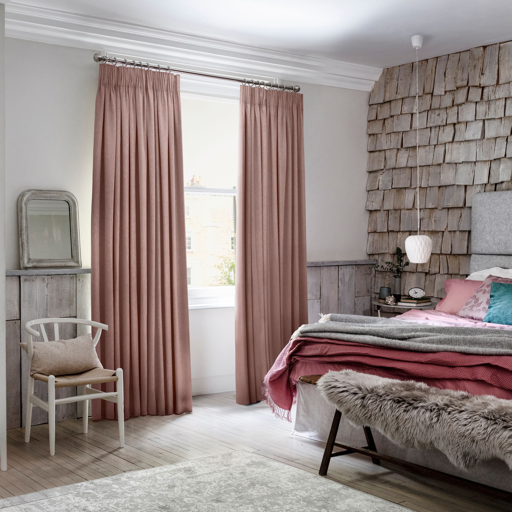 A cozy bedroom features pink curtains covering a window, beside a wooden chair with a cushion. A bed with layered blankets and a fur-covered bench sits near a shingled accent wall.
