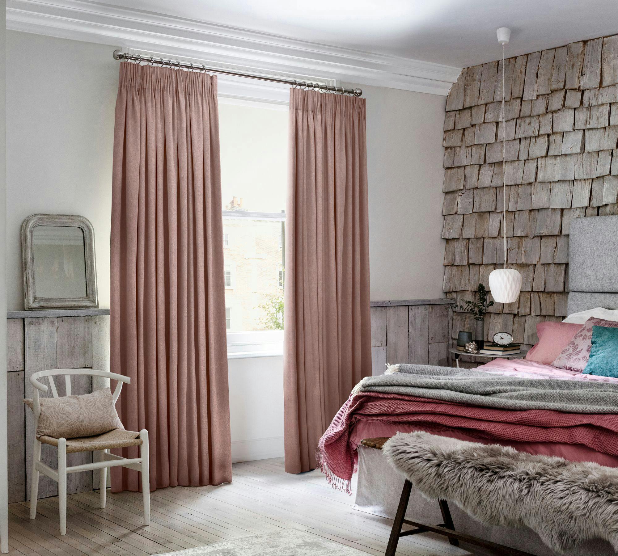 A cozy bedroom features pink curtains covering a window, beside a wooden chair with a cushion. A bed with layered blankets and a fur-covered bench sits near a shingled accent wall.