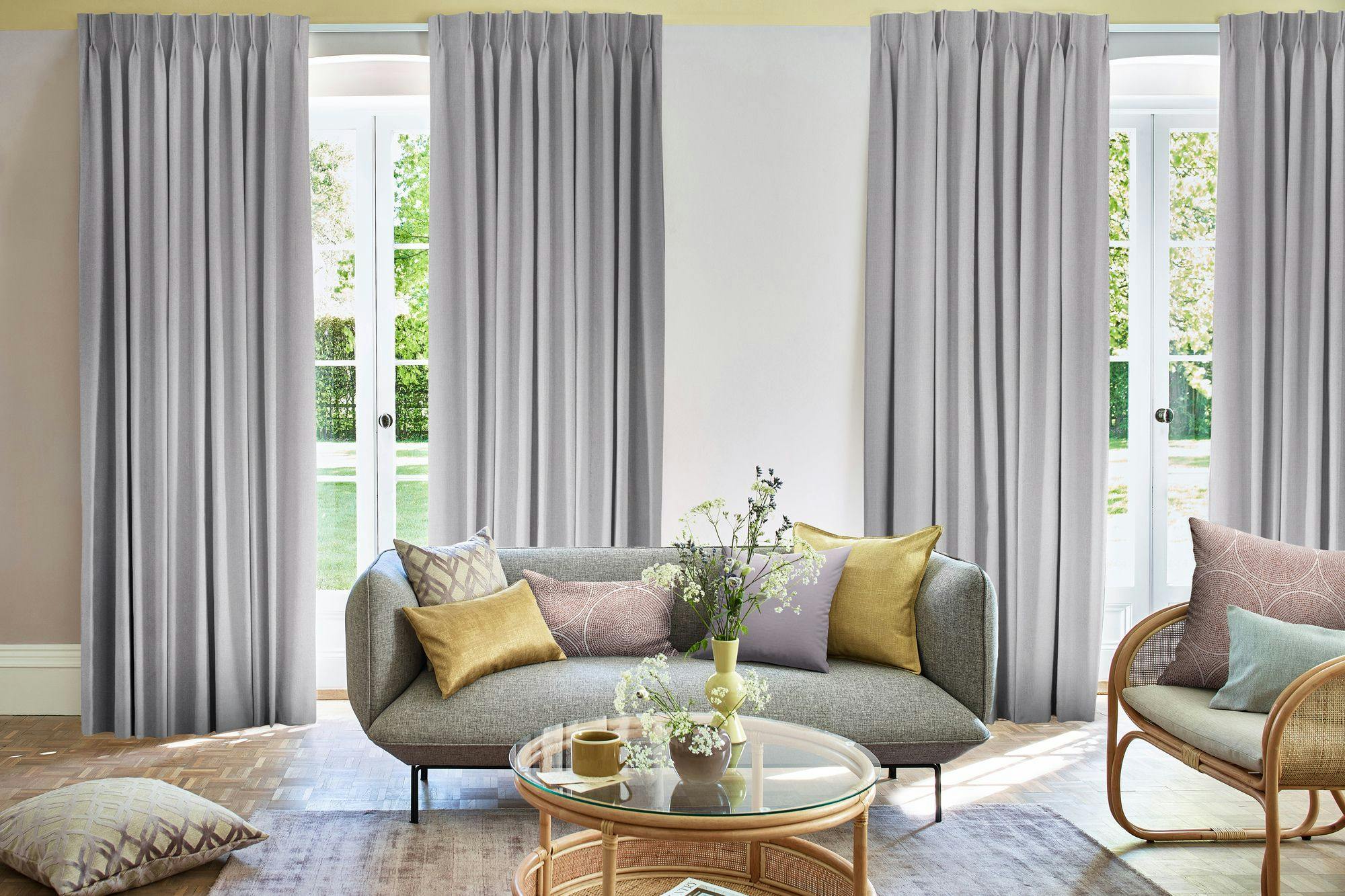 A gray sofa with colorful cushions sits beside a glass coffee table with flowers, set in a room with white curtains and large windows overlooking a green garden.