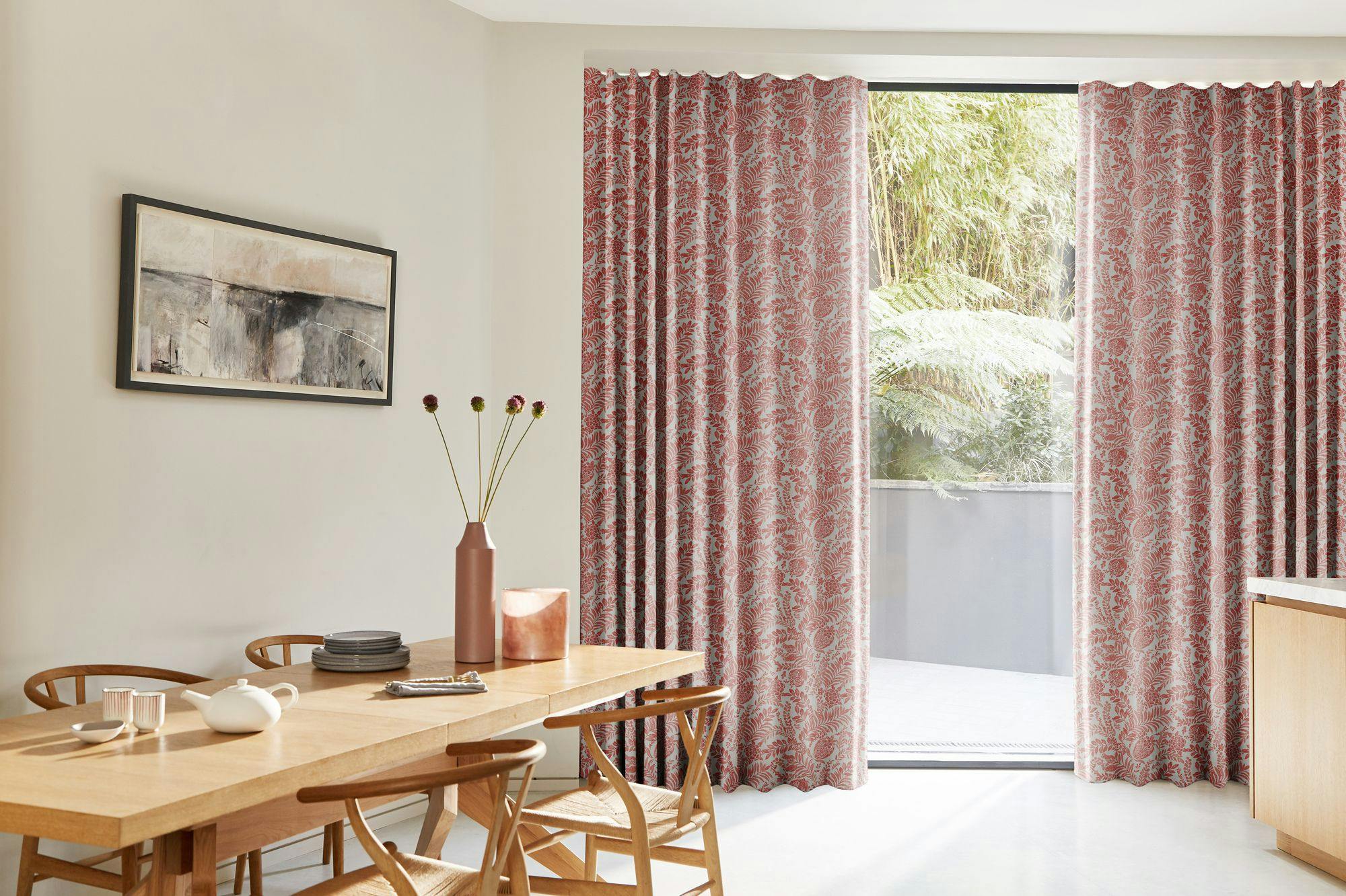 Dining table holds a teapot, bowls, and vase, positioned beside red-patterned curtains open to a view of lush greenery. Wall art hangs above wooden chairs in a bright room.
