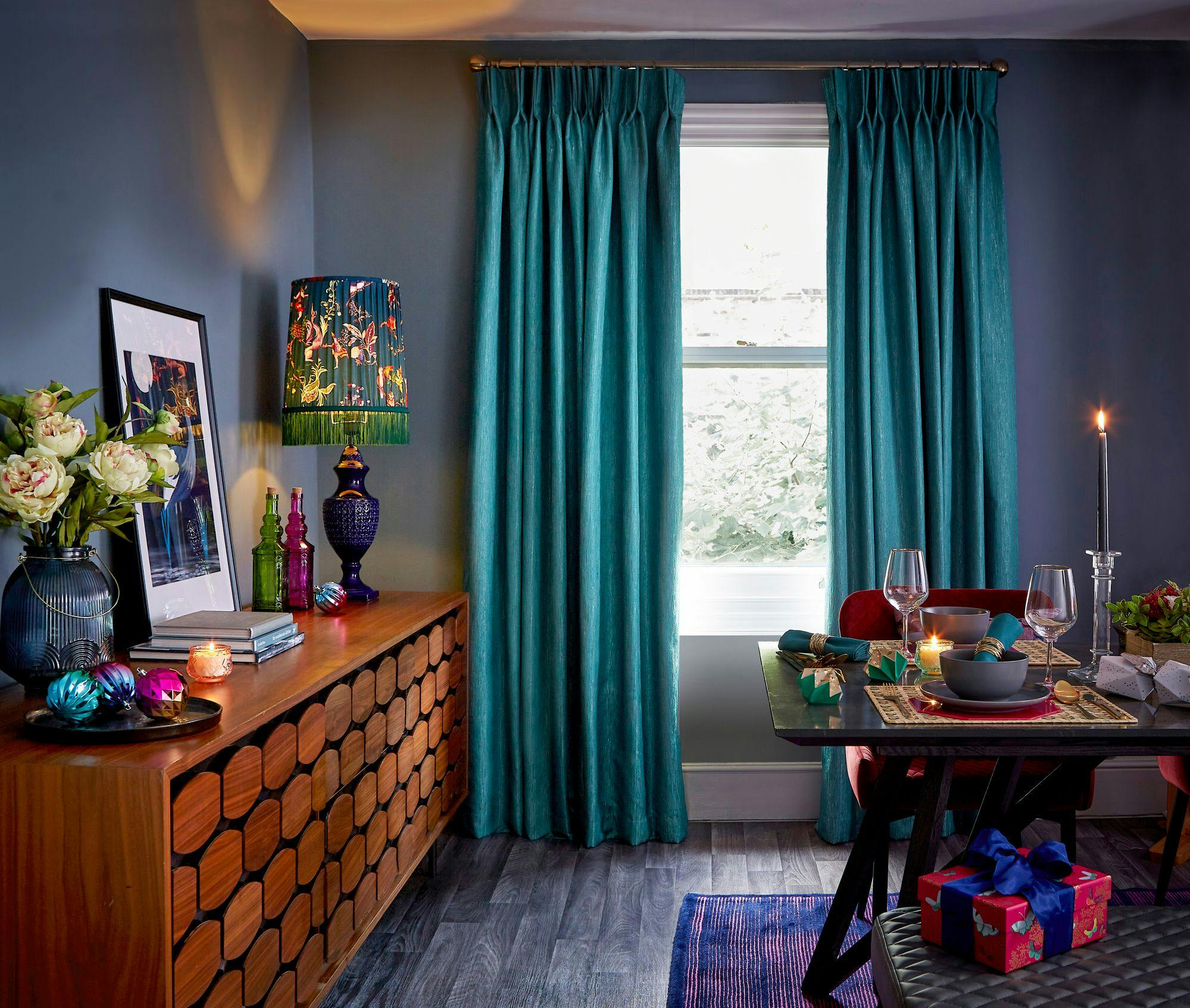 Sideboard holding books, flowers, and a decorative lamp illuminates, next to a window with teal curtains. Dining table set elegantly, includes candles and a gift-wrapped box, enhancing cozy ambience.