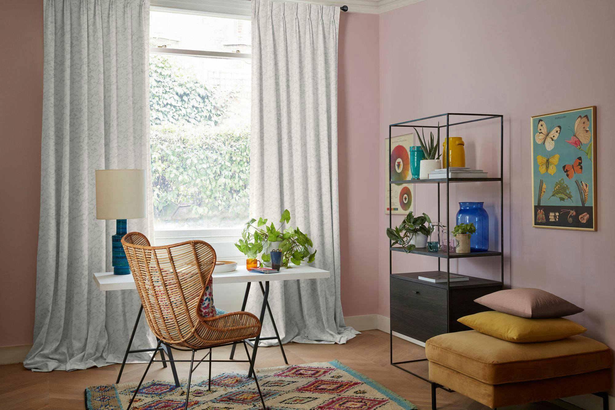 A wicker chair faces a white desk with plants and a lamp, in a room with pink walls and a window with curtains. Nearby, a shelving unit holds books, colorful vases, and more plants.