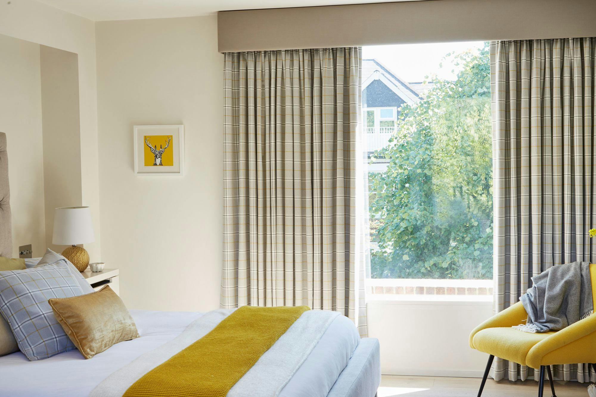 Bed with yellow blanket and pillows, next to a nightstand with a lamp, faces a window with plaid curtains. A framed deer illustration hangs on a cream wall.