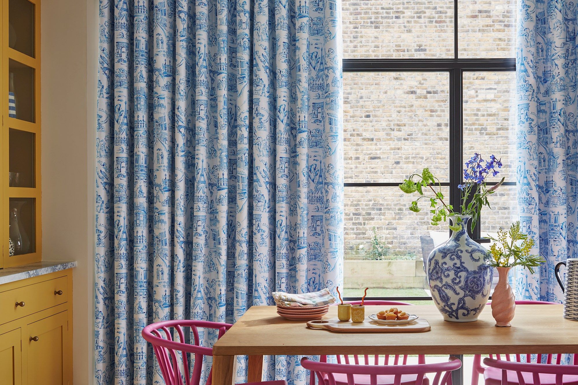 Curtains with blue intricate patterns hang beside a large window overlooking a brick wall, while a dining table adorned with a floral vase, plates, and food sits underneath.