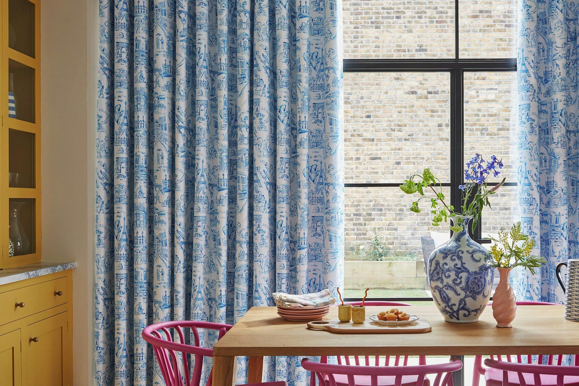 Curtains with blue intricate patterns hang beside a large window overlooking a brick wall, while a dining table adorned with a floral vase, plates, and food sits underneath.