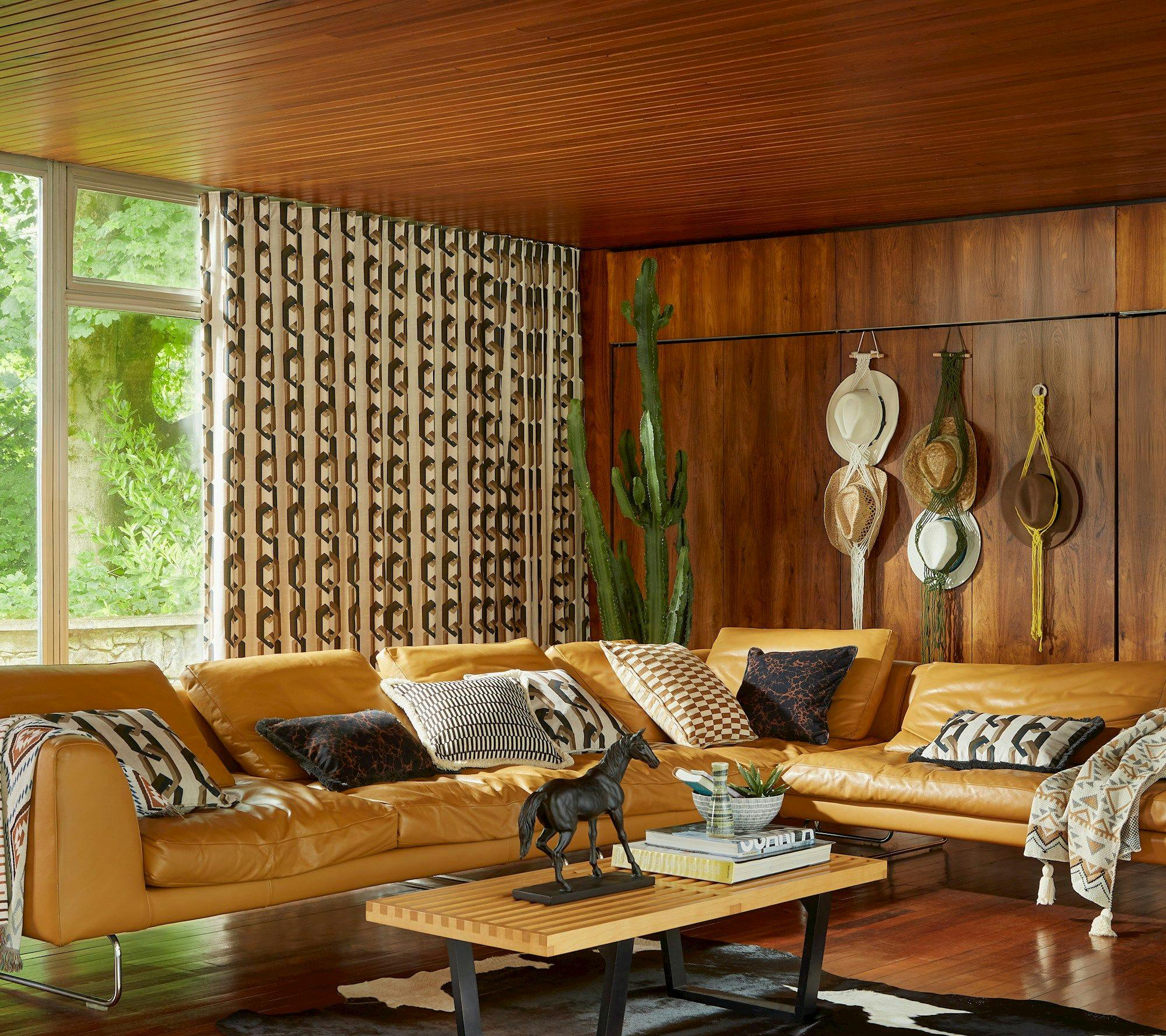 A cozy living room features a mustard-colored sofa adorned with patterned cushions, set against a wood-paneled wall with hanging hats. A horse statue sits on the coffee table, and a cactus adds greenery.