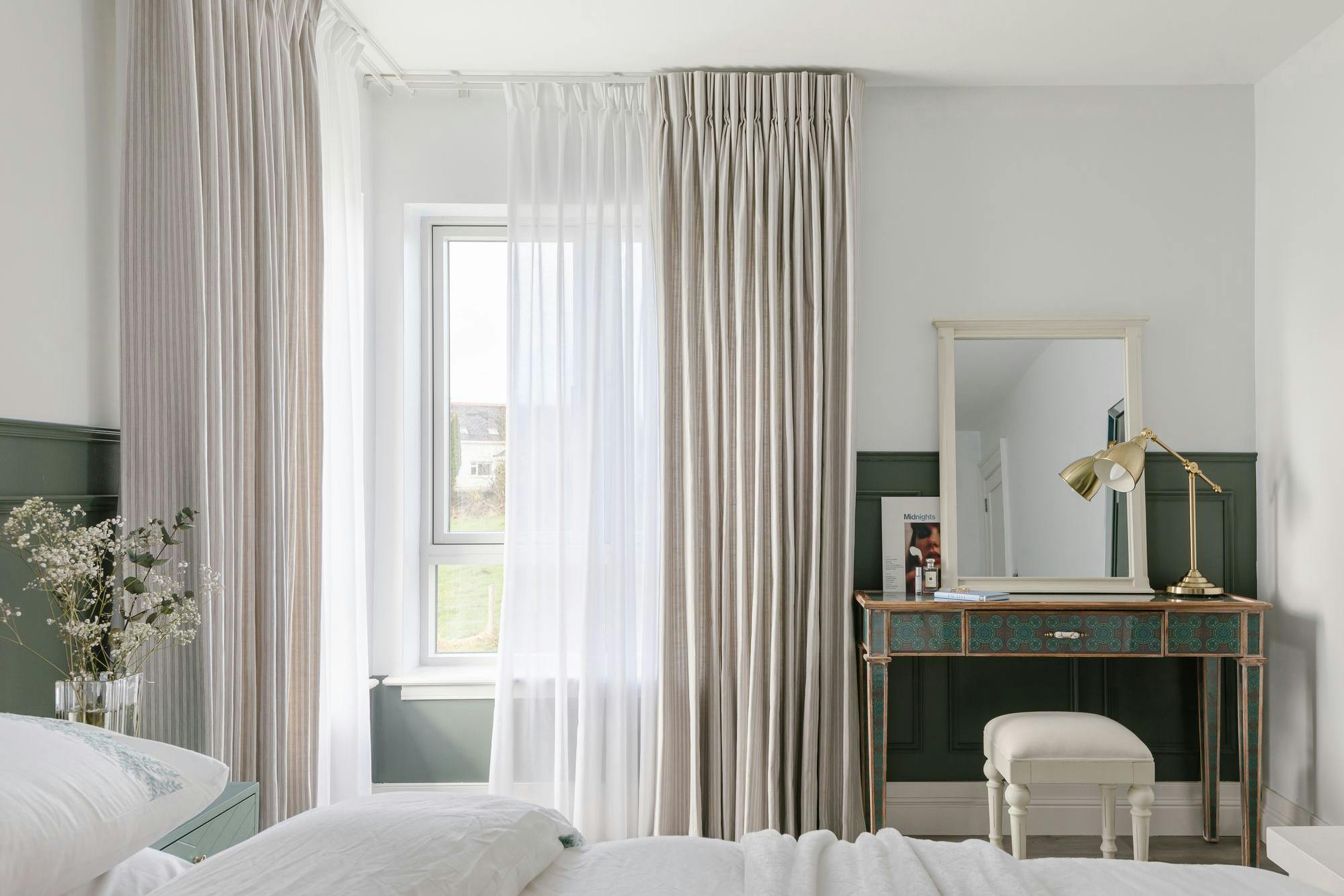 A bedroom features a bed with white bedding, a window with sheer and striped curtains, a vintage-style desk with a stool, a gold lamp, and a small mirror.
