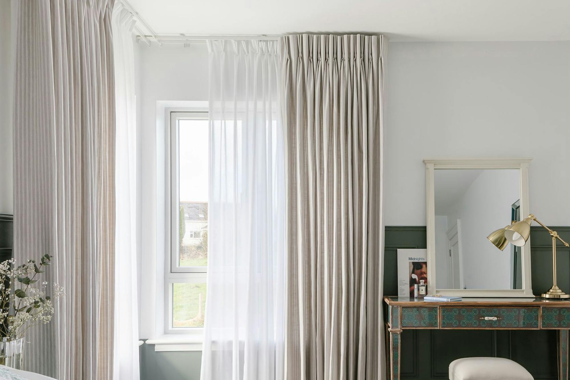 A bedroom features a bed with white bedding, a window with sheer and striped curtains, a vintage-style desk with a stool, a gold lamp, and a small mirror.