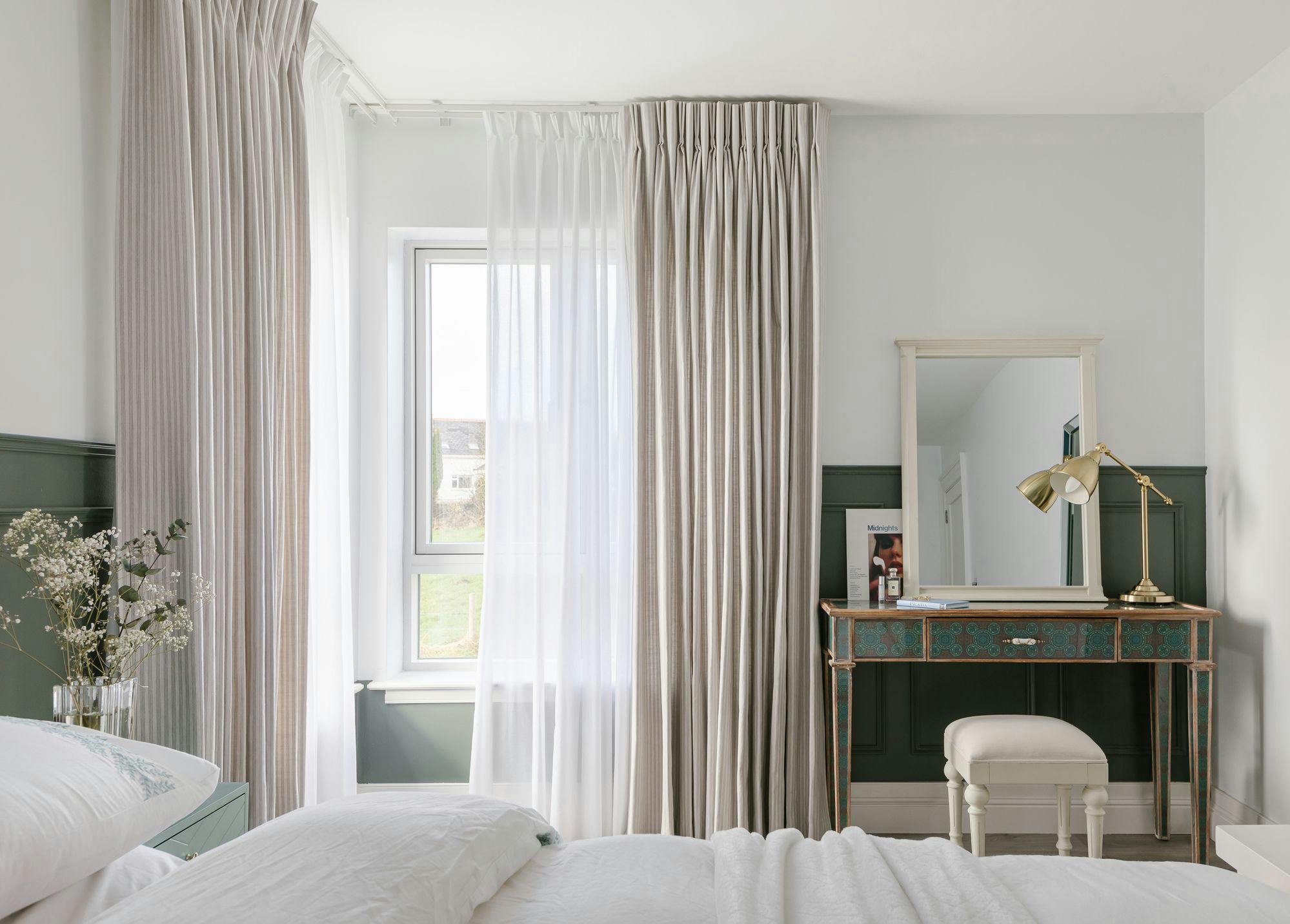 A bedroom features a bed with white bedding, a window with sheer and striped curtains, a vintage-style desk with a stool, a gold lamp, and a small mirror.