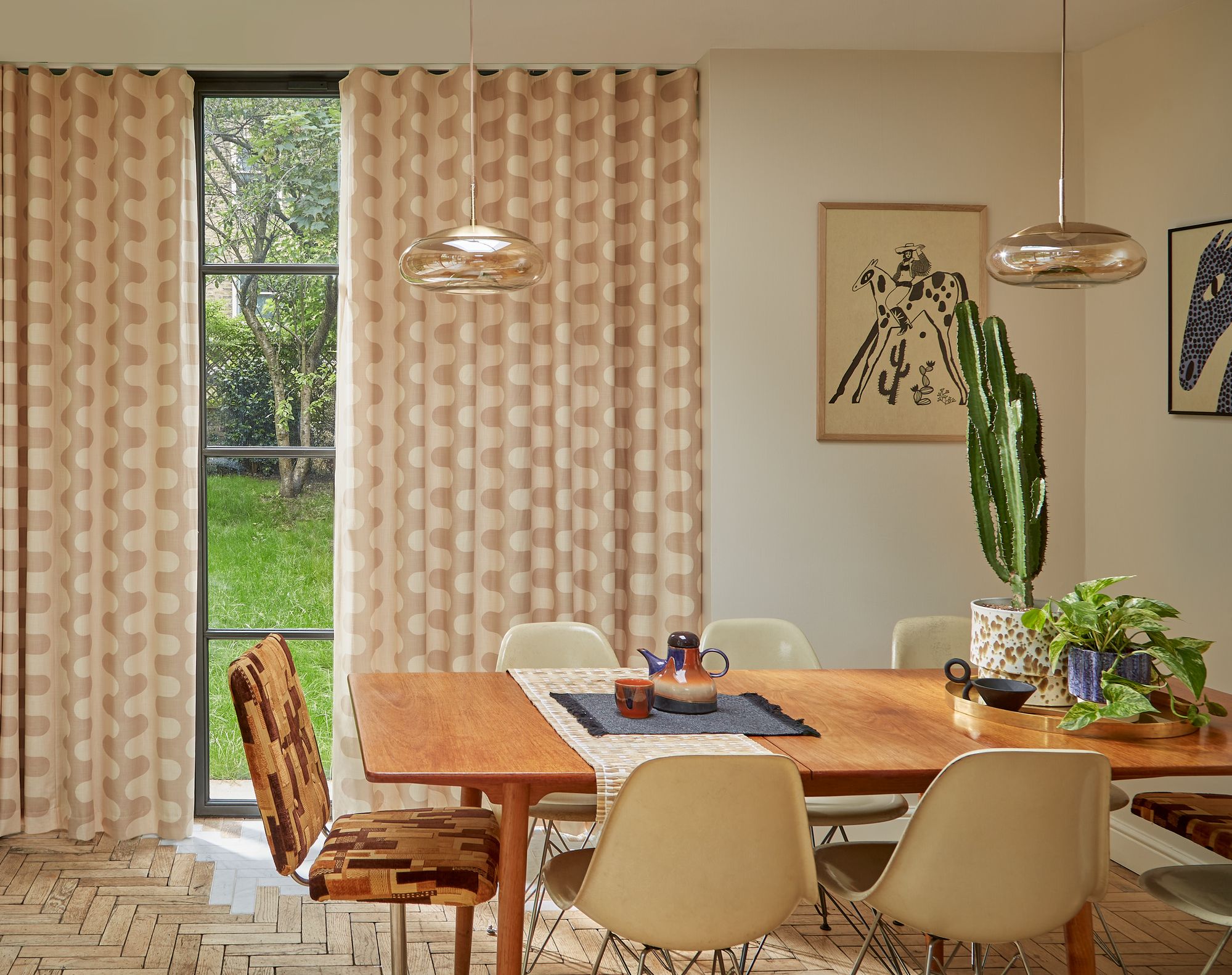 Dining table set with chairs surrounding it, featuring a cactus and plants on top, situated in a room with patterned curtains, artwork on walls, and a large window showing green grass outside.