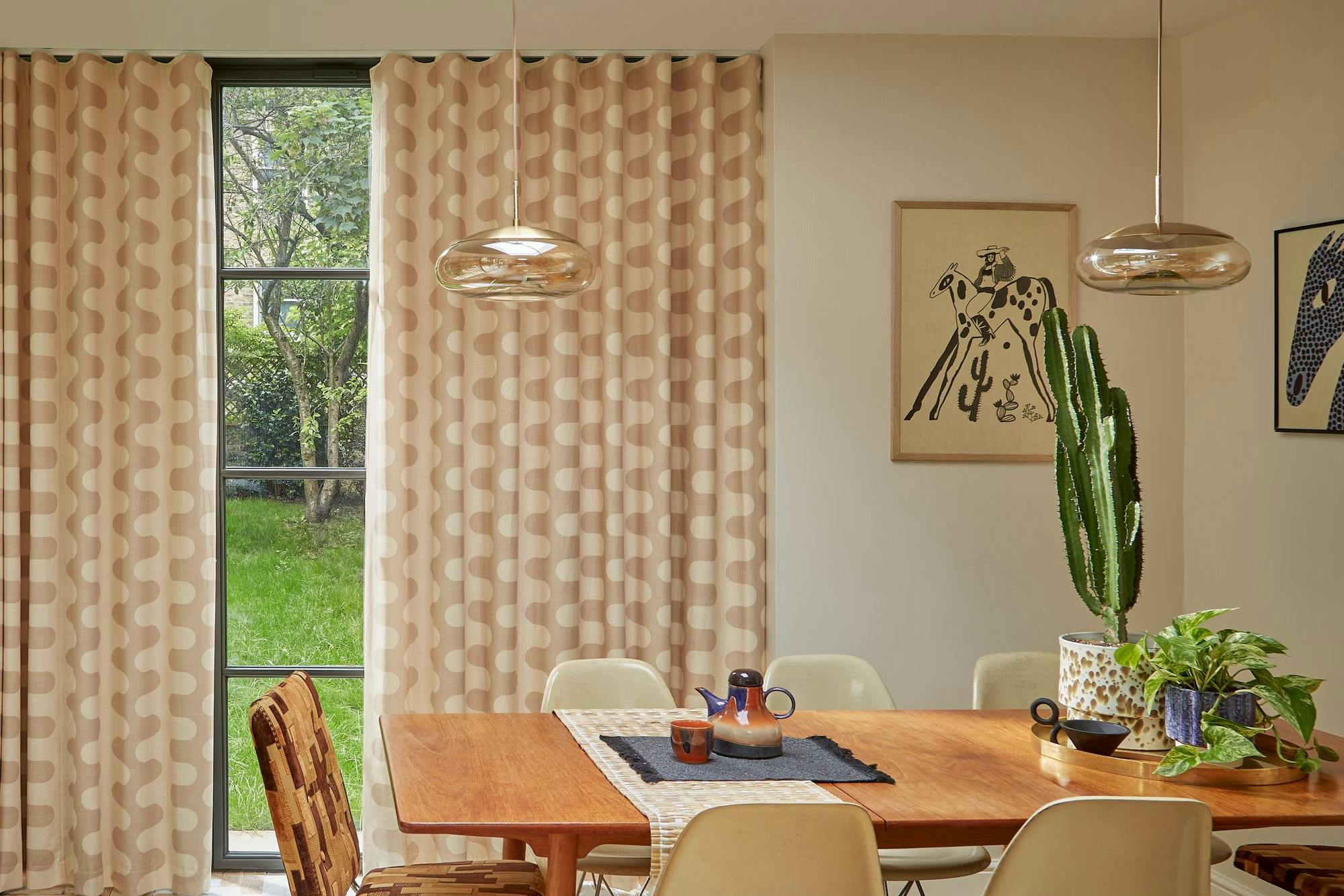 Dining table set with chairs surrounding it, featuring a cactus and plants on top, situated in a room with patterned curtains, artwork on walls, and a large window showing green grass outside.