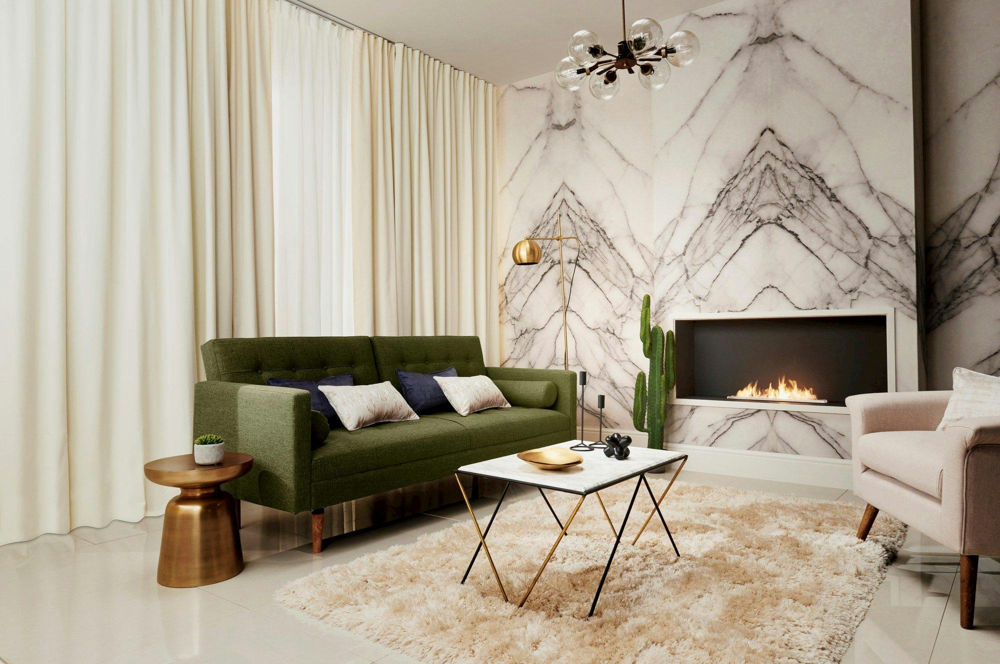 A green sofa with cushions sits in a stylish living room, next to a cozy fire in a modern fireplace. A marble-patterned wall, beige curtains, and shaggy rug complete the elegant space.