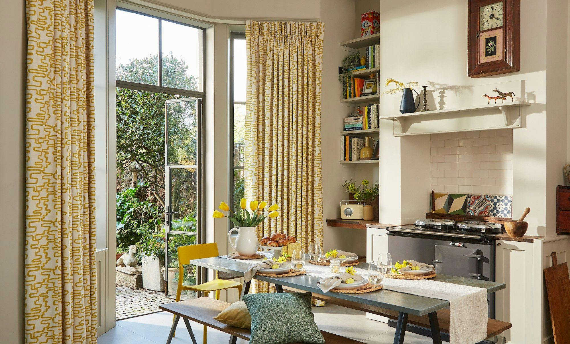 A dining table set with plates, glasses, and a vase of yellow tulips sits near a sunny window with open glass doors, revealing a lush garden outside.