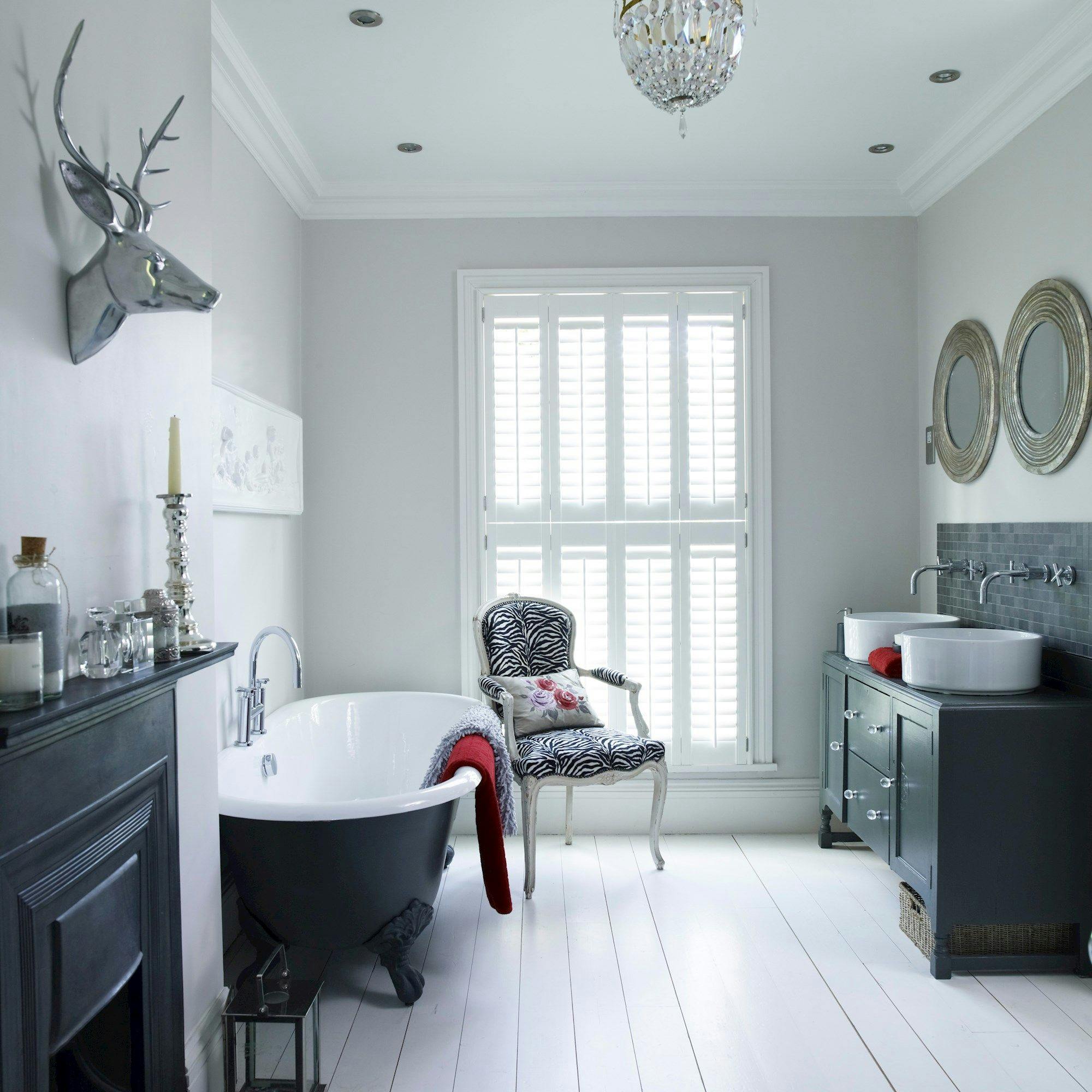 Clawfoot bathtub with red towel sits beside black fireplace, decorative stag head, and silver candlestick. Nearby, a zebra-print chair faces two sinks under round mirrors, all illuminated by chandelier.