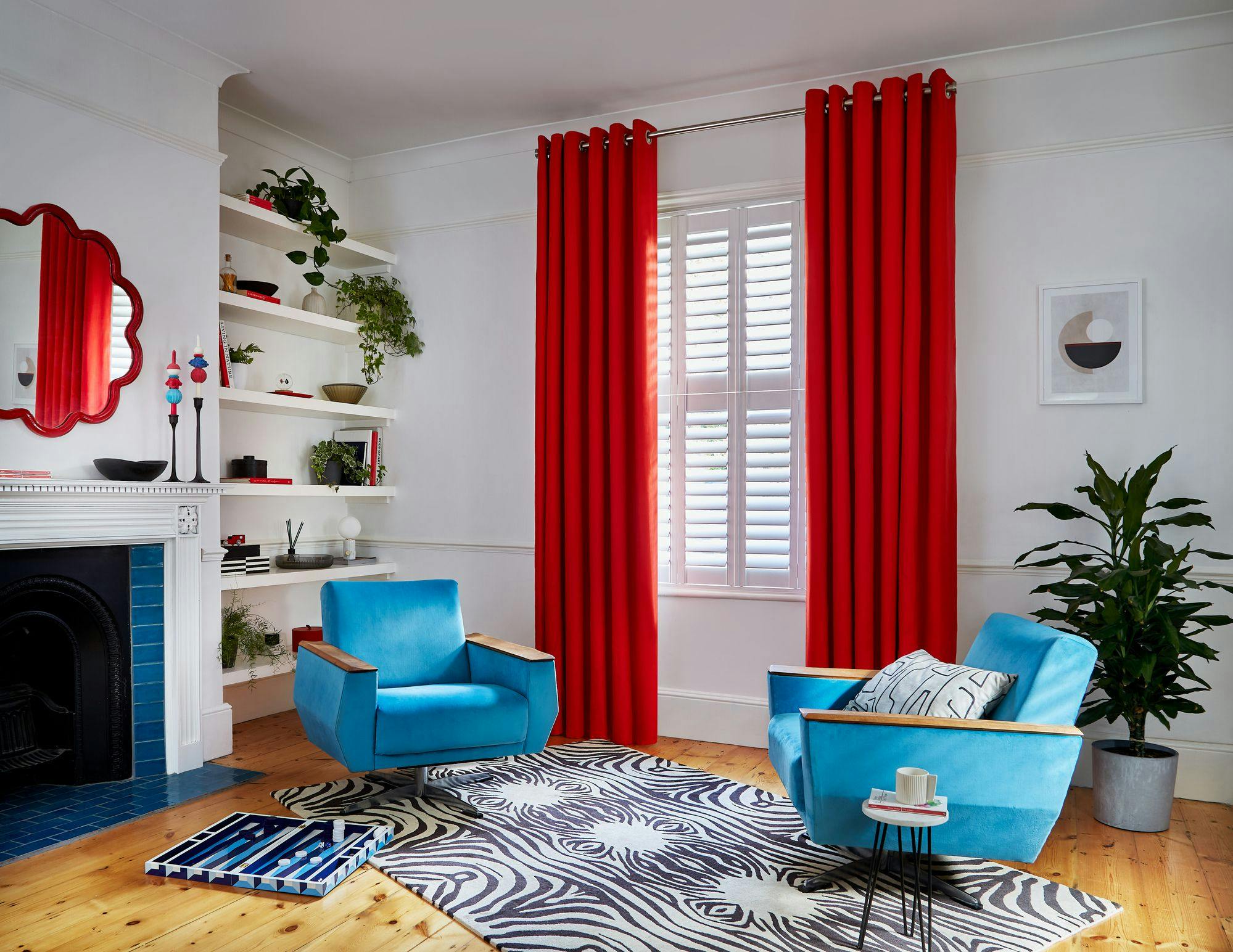 Two blue armchairs face each other on a zebra-patterned rug in a cozy living room, surrounded by red curtains, shelves with decor, a fireplace with a mirror, and a plant.