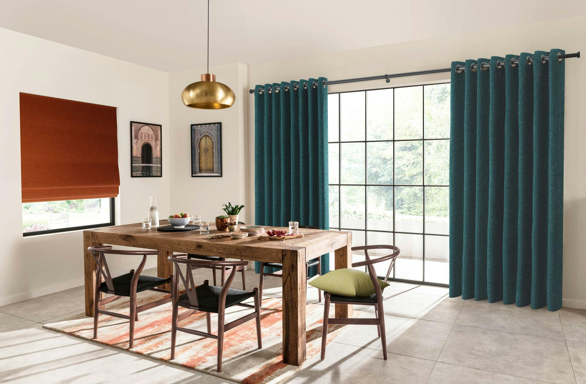 A wooden dining table with chairs sits under a gold pendant light. Blue curtains frame large windows. Red blinds cover another window. Two framed artworks decorate the cream walls.