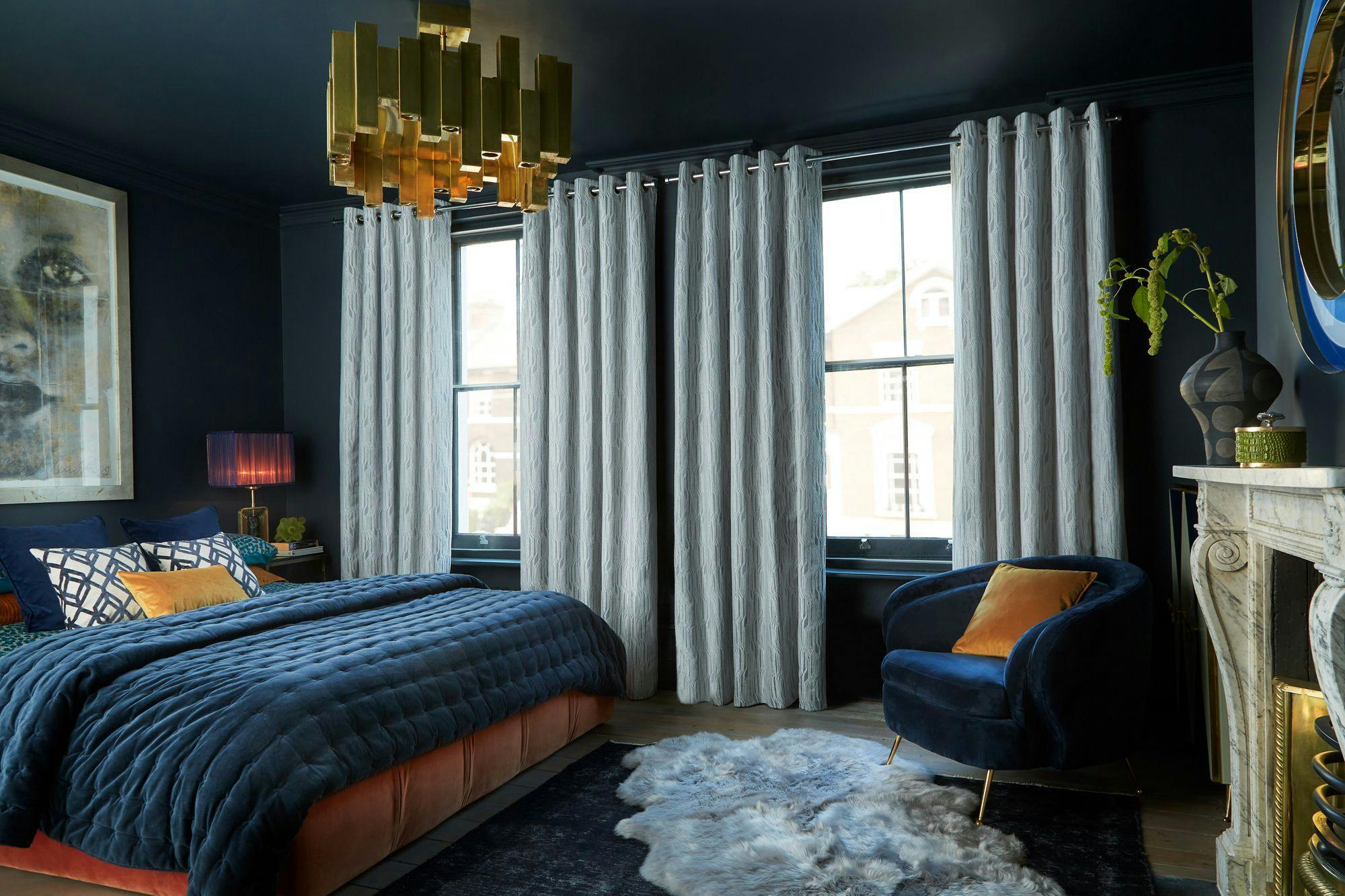 A bedroom features a navy blue velvet bed, accented cushions, and a plush armchair. Gold chandelier overhead, large windows with white curtains, rug, and ornate fireplace complete the elegant decor.