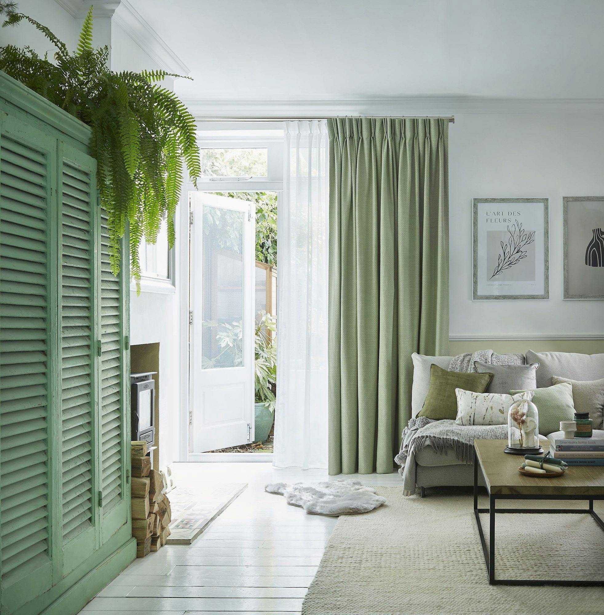 A cozy living room features a white sofa with green cushions, a wooden coffee table, and lush plants atop a green cabinet. Sunlight streams through open double doors beside green curtains.