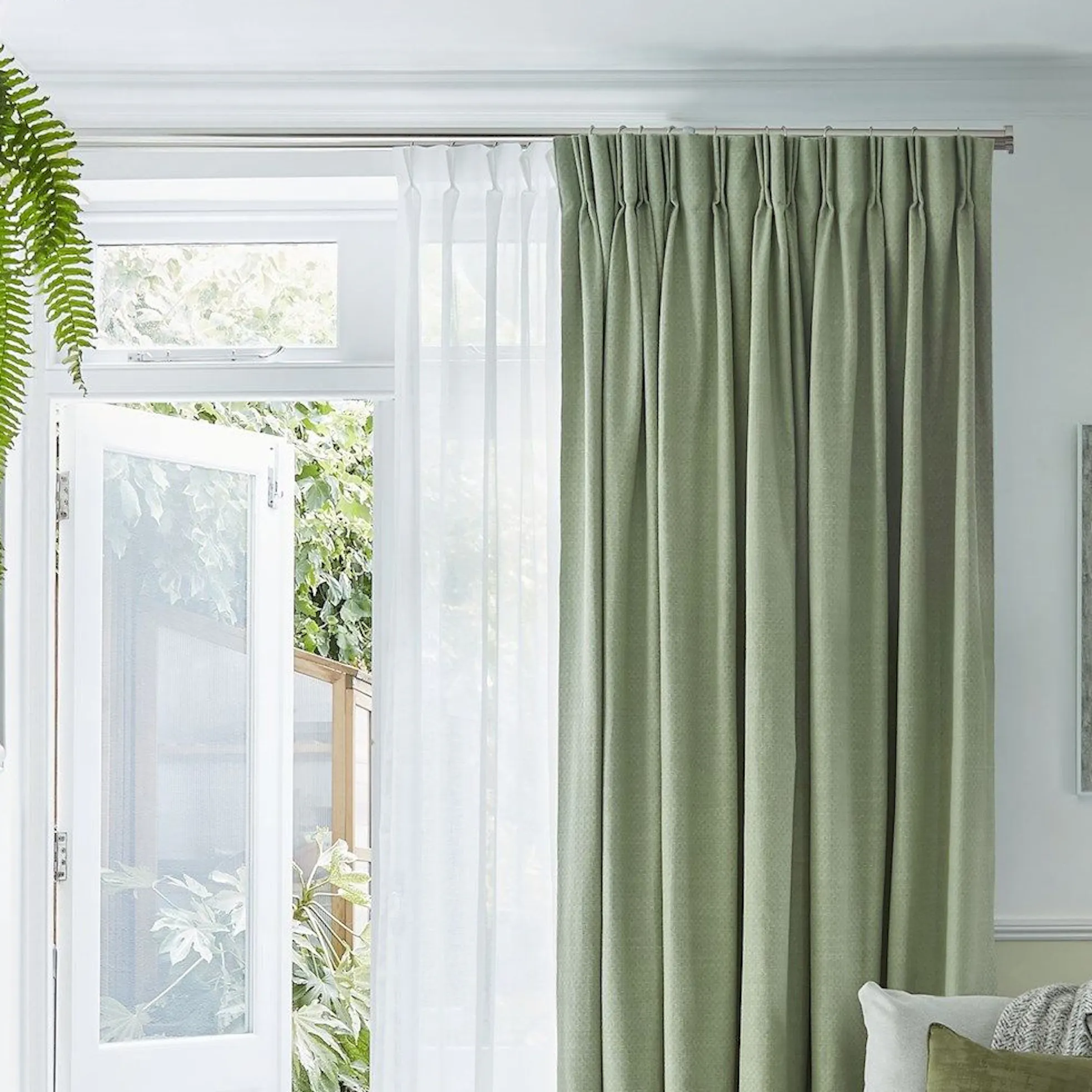 A cozy living room features a white sofa with green cushions, a wooden coffee table, and lush plants atop a green cabinet. Sunlight streams through open double doors beside green curtains.
