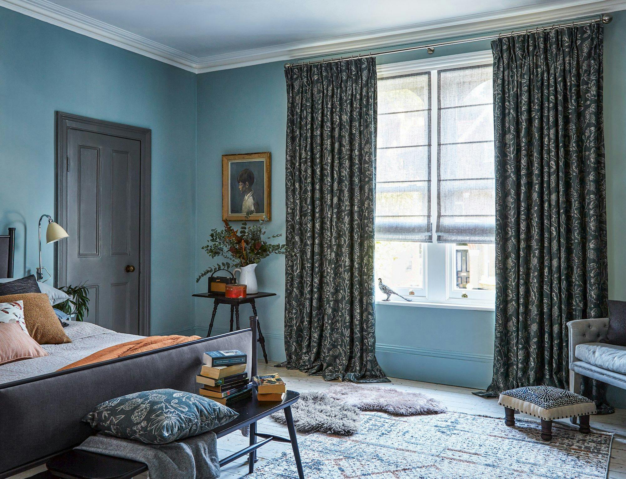 A cozy bedroom features a neatly made bed, books stacked on a bench, and a side table with a vase of flowers. Curtains frame a window, brightening the blue-painted room.