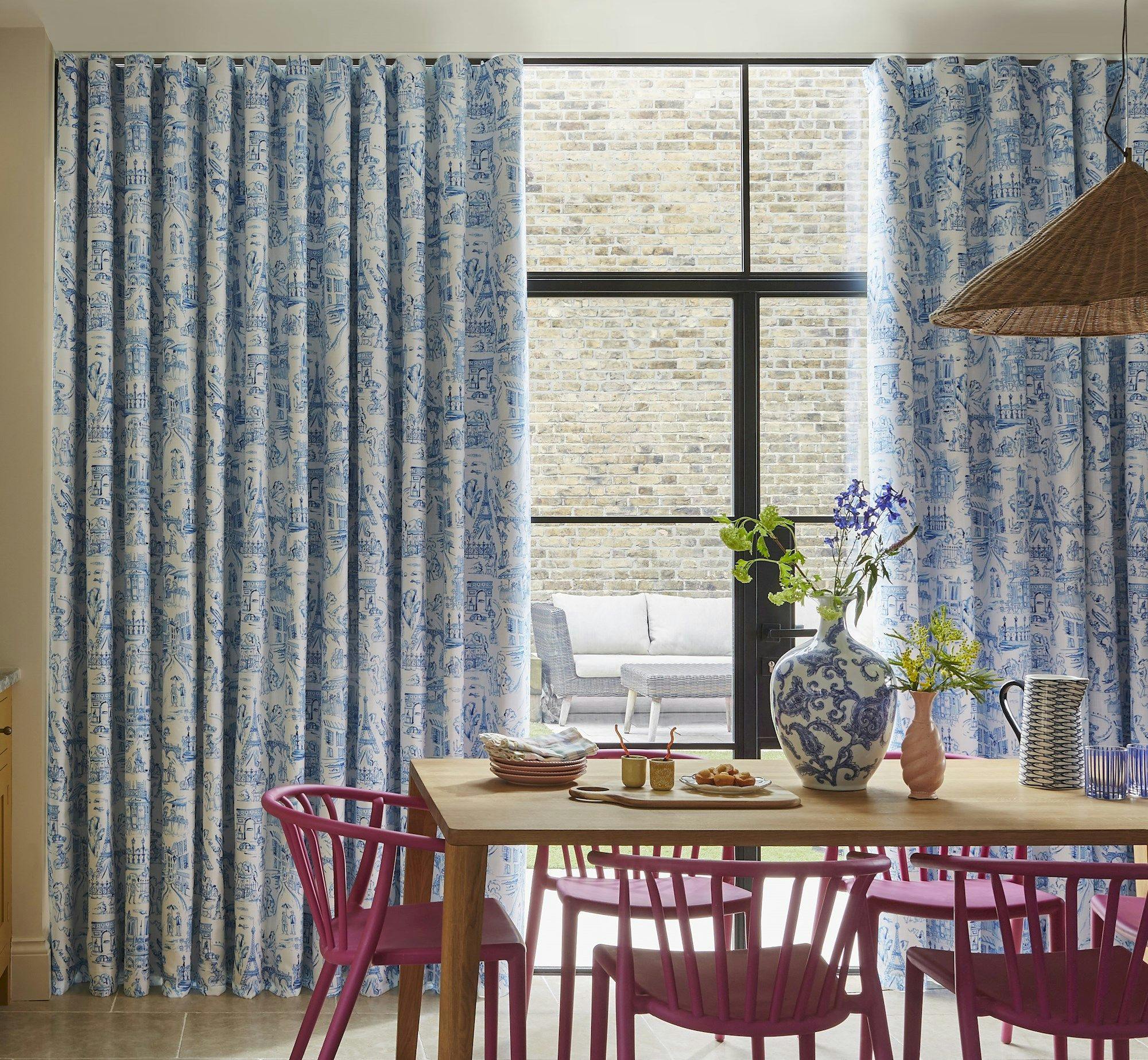 Curtains with blue pattern hang beside a large window. A wooden table with purple chairs displays a vase with flowers, plates, and cups. A brick wall and outdoor seating are visible outside.