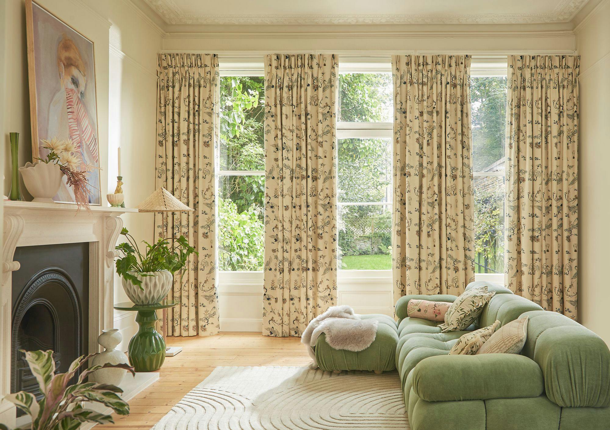 A plush green sofa with cushions sits on a patterned rug in a bright room. Large windows with floral curtains overlook a lush garden. Art decorates the walls.
