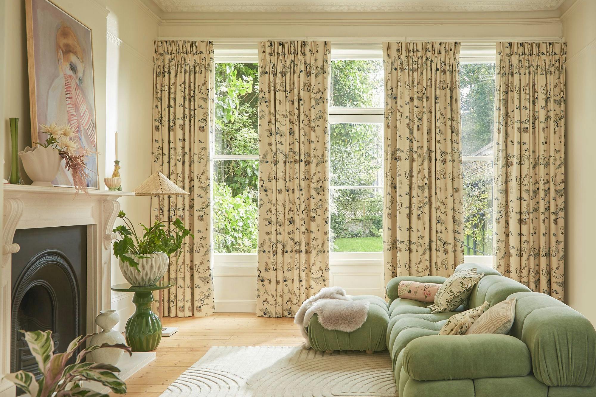 A plush green sofa with cushions sits on a patterned rug in a bright room. Large windows with floral curtains overlook a lush garden. Art decorates the walls.