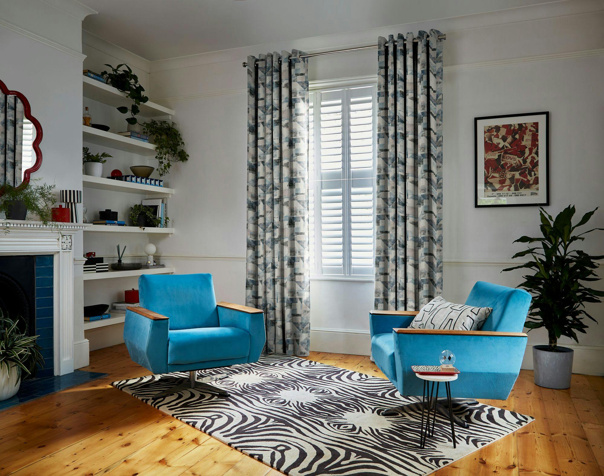 Two blue armchairs sit on a zebra-patterned rug in a living room with wooden flooring, surrounded by shelves, plants, a fireplace, and patterned curtains framing a window.
