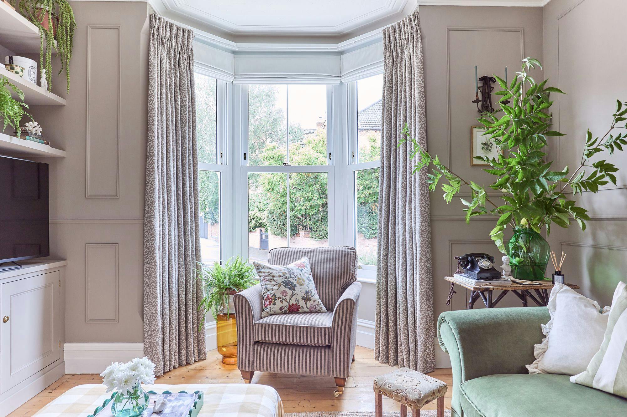 An armchair with a floral cushion sits by a large bay window, framed by patterned curtains. Surrounding are indoor plants, a small table with decor, and a green sofa in a cozy room.