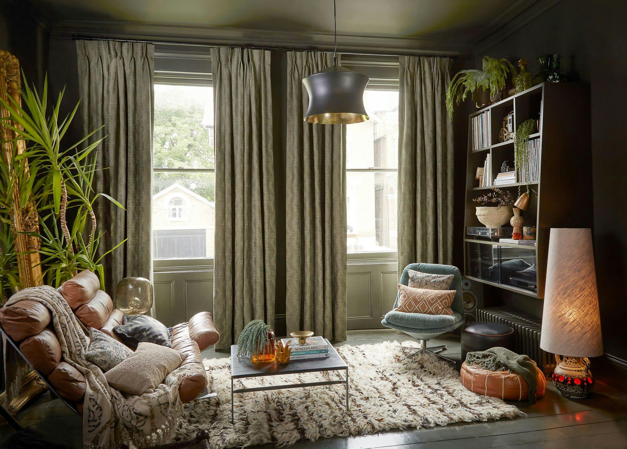 A cozy living room features a brown leather sofa with cushions, a central coffee table holding books and amber bottles, and a large window framed by curtains. A lamp softly lights nearby shelves.