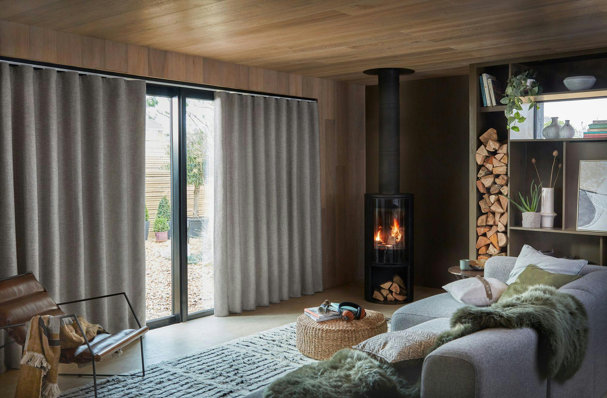 A cozy living room features a crackling fire in a black stove, surrounded by logs and bookshelves. A gray sofa with cushions faces floor-length windows covered by beige curtains.