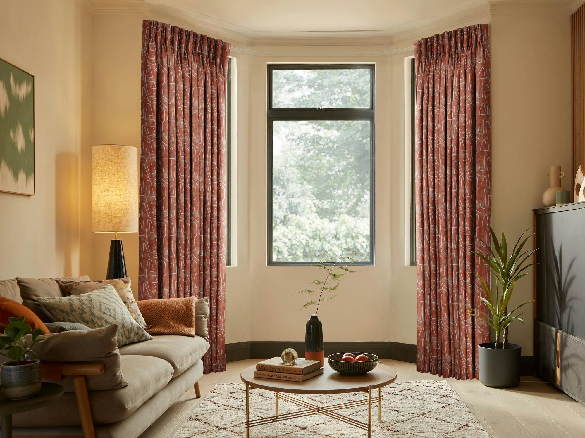 Curtains hang beside a large window, framing a view of greenery. A cozy living room features a beige sofa, modern floor lamp, and circular wooden coffee table on a patterned rug.