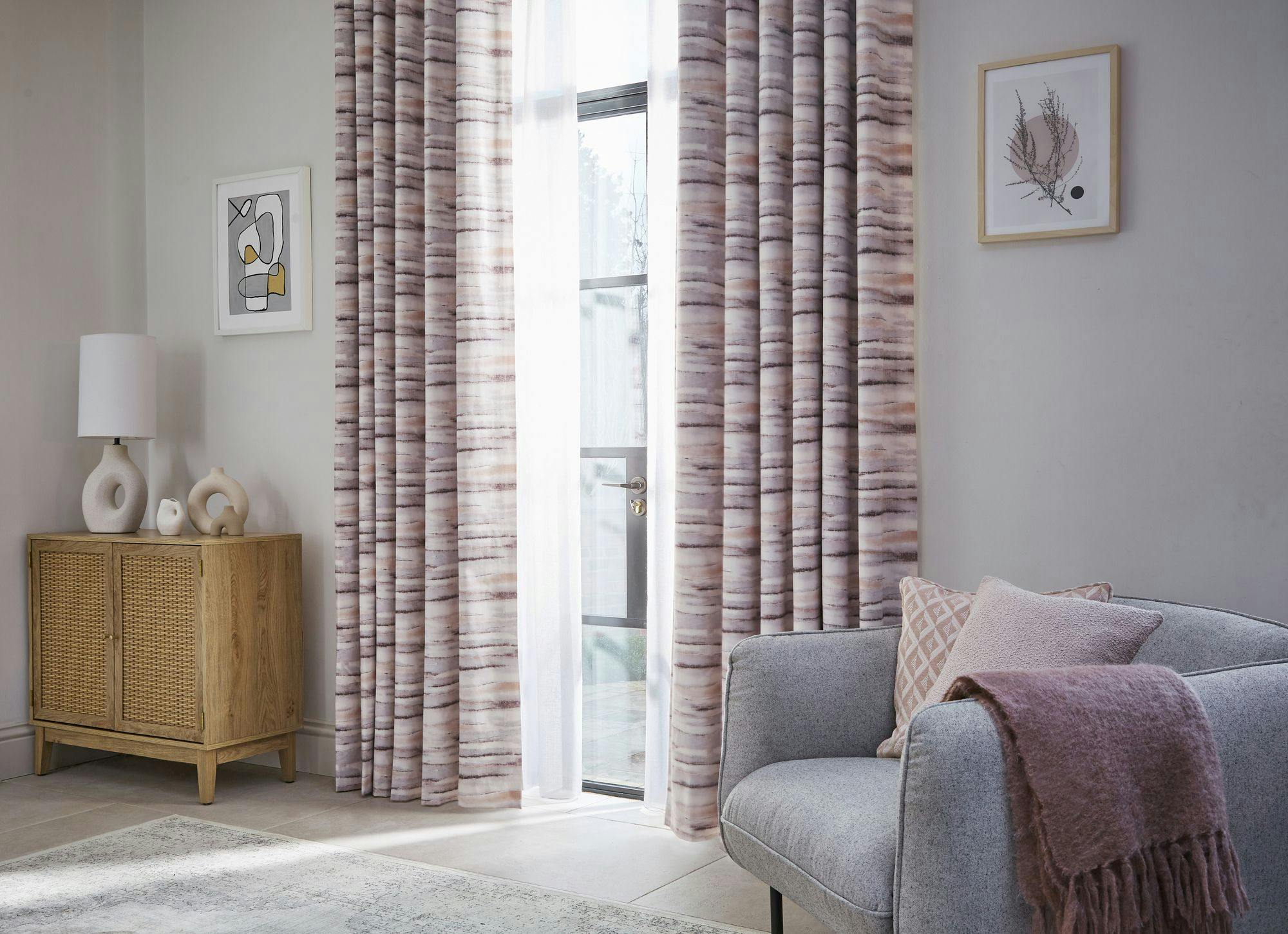 Curtains hang beside a tall window, framing a cozy living room with a gray sofa, wooden cabinet, and two framed artworks. Soft pillows and a throw blanket add warmth.